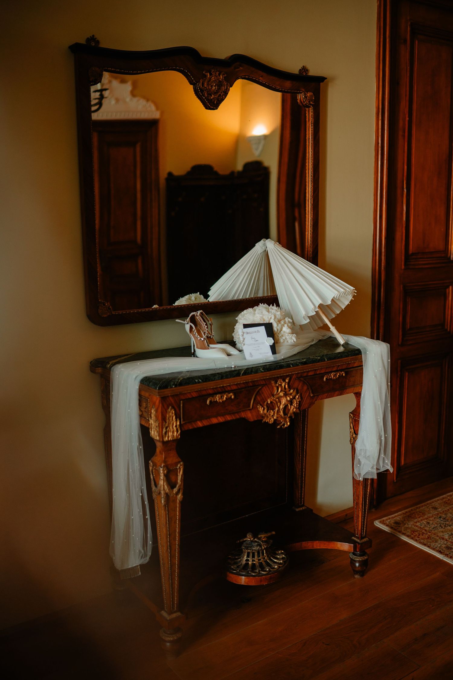 Antique wooden vanity with ornate mirror, white parasol, and draped fabric in warm interior.