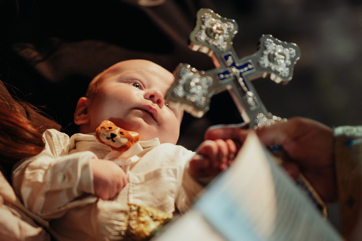 A baby in white christening gown looks up as a silver cross is held nearby.