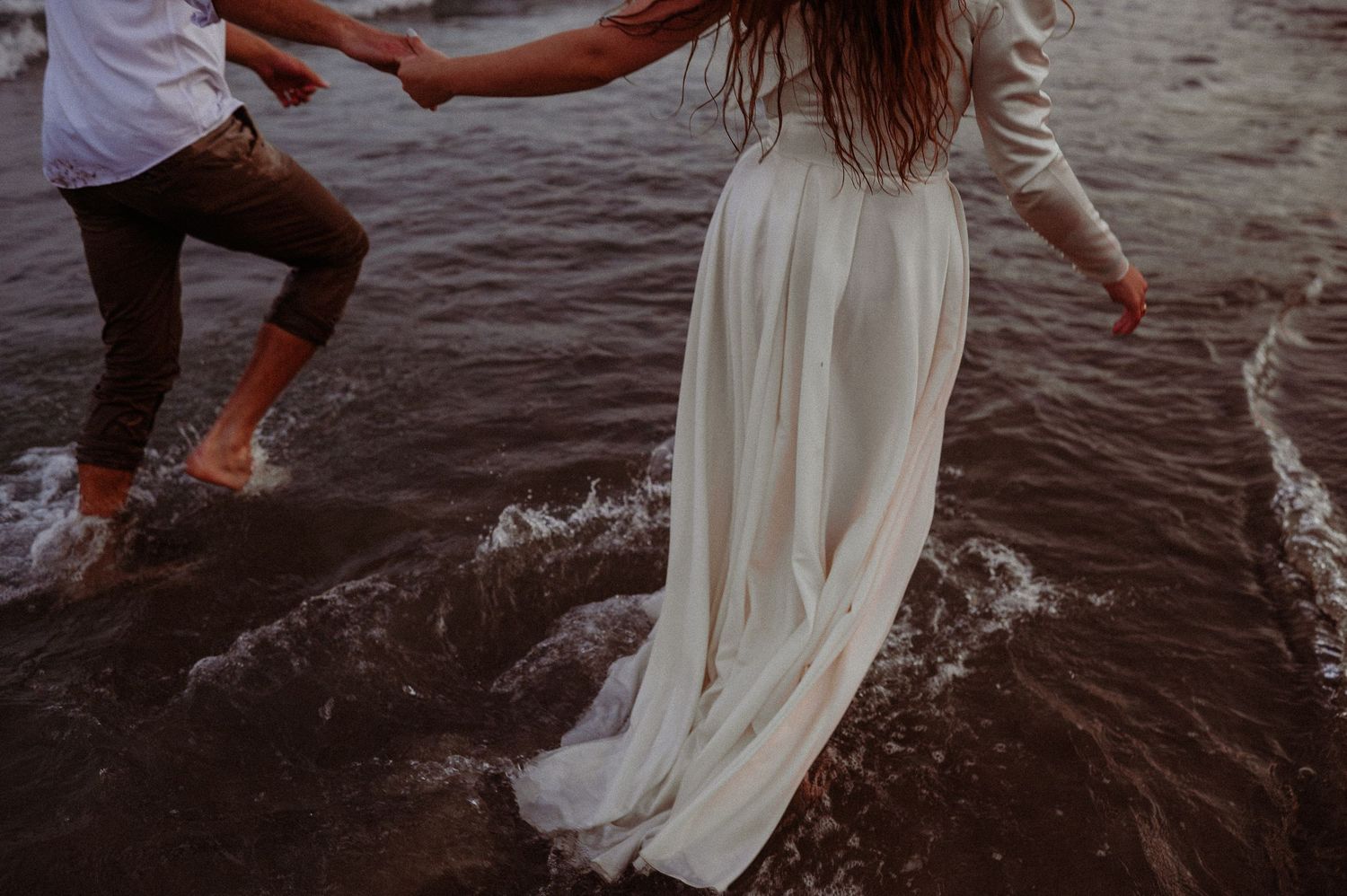 Couple holding hands and running through shallow ocean waves, woman wearing a flowing white dress.