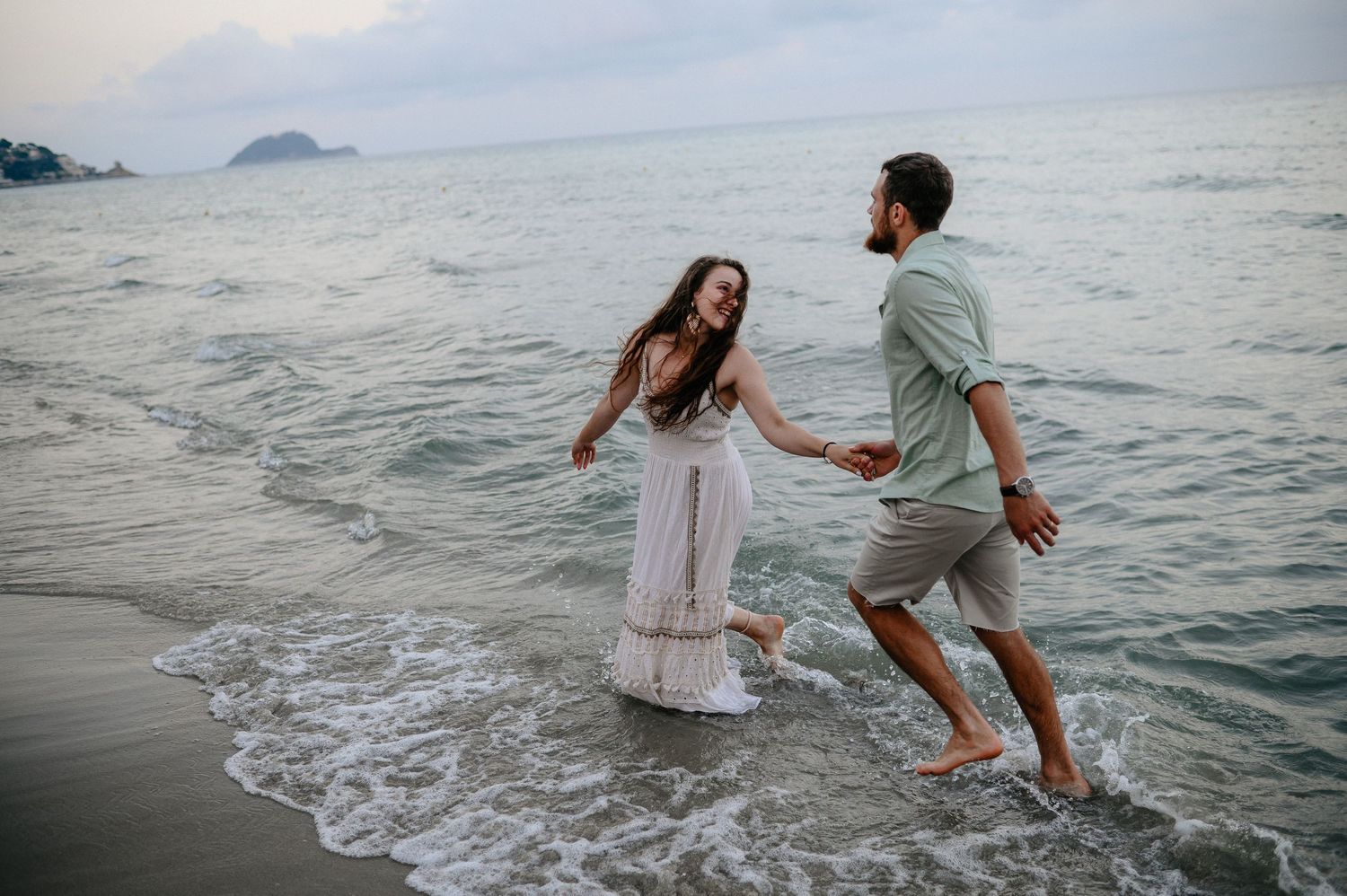 Playful couple holding hands and splashing through ocean waves on a cloudy beach with rocky islands.