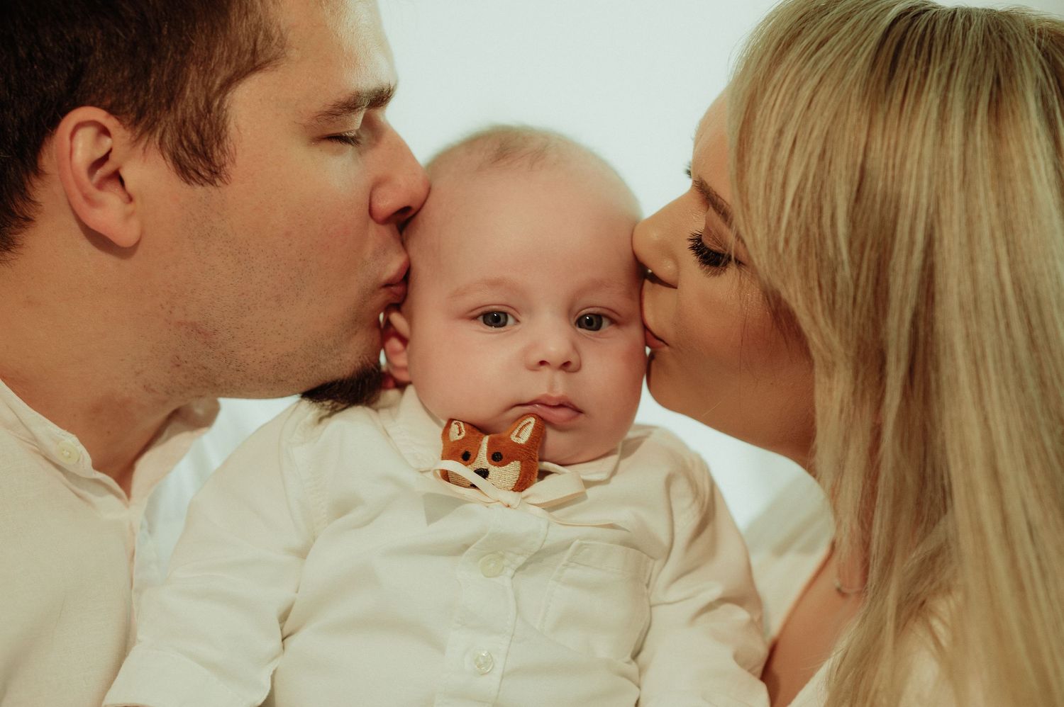 Two parents kissing their baby on both cheeks simultaneously in a sweet family portrait.