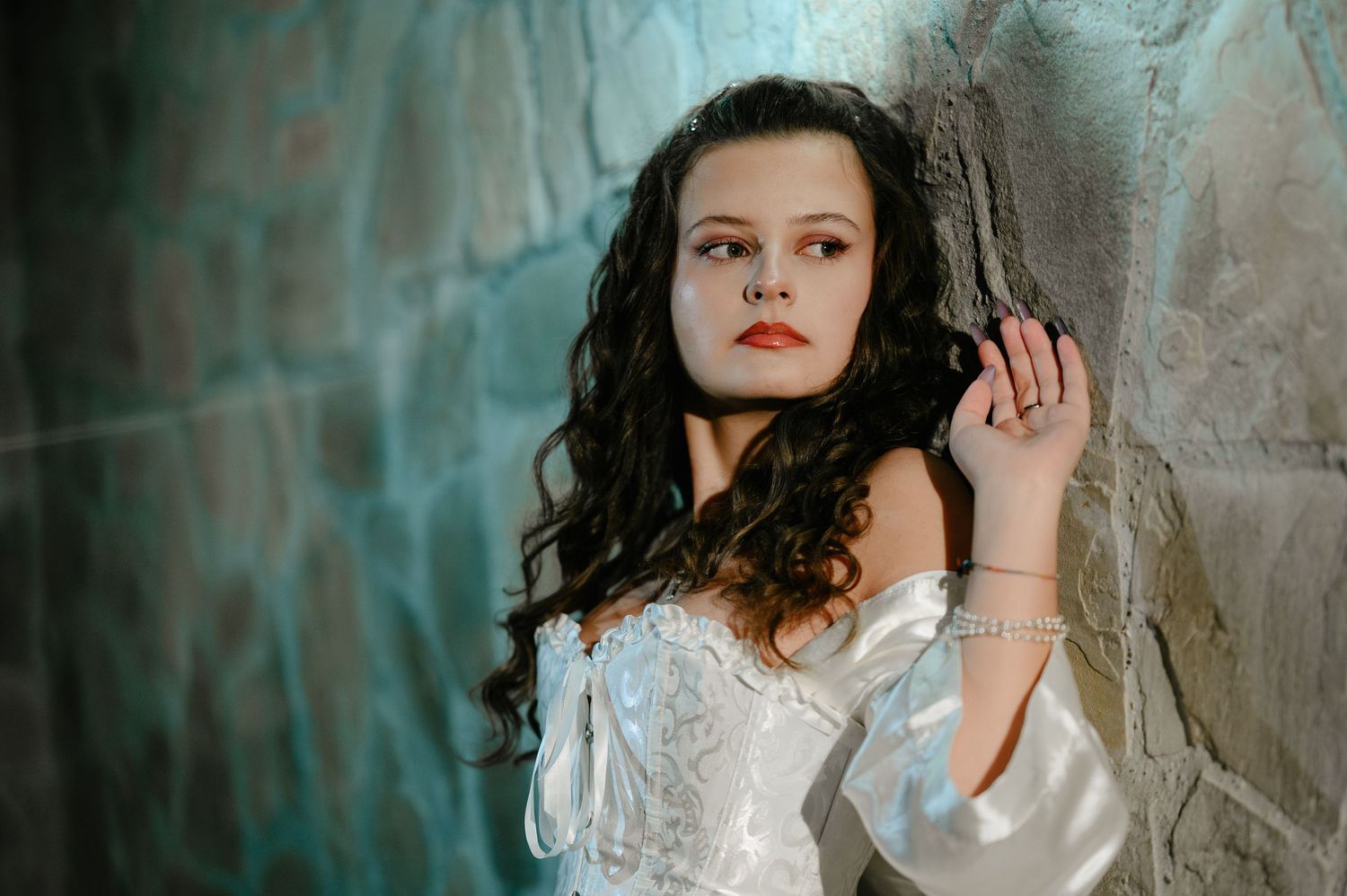 Young woman with curly hair in white corset dress leans against stone wall in moody portrait.