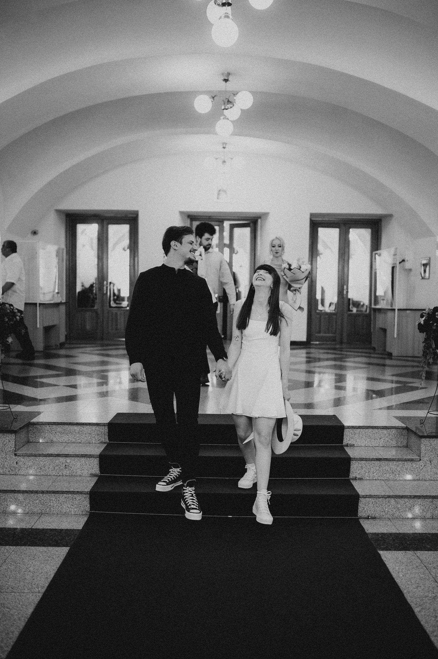 Black and white photo of newlyweds walking down red carpet in grand hall, followed by wedding guests carrying flowers.