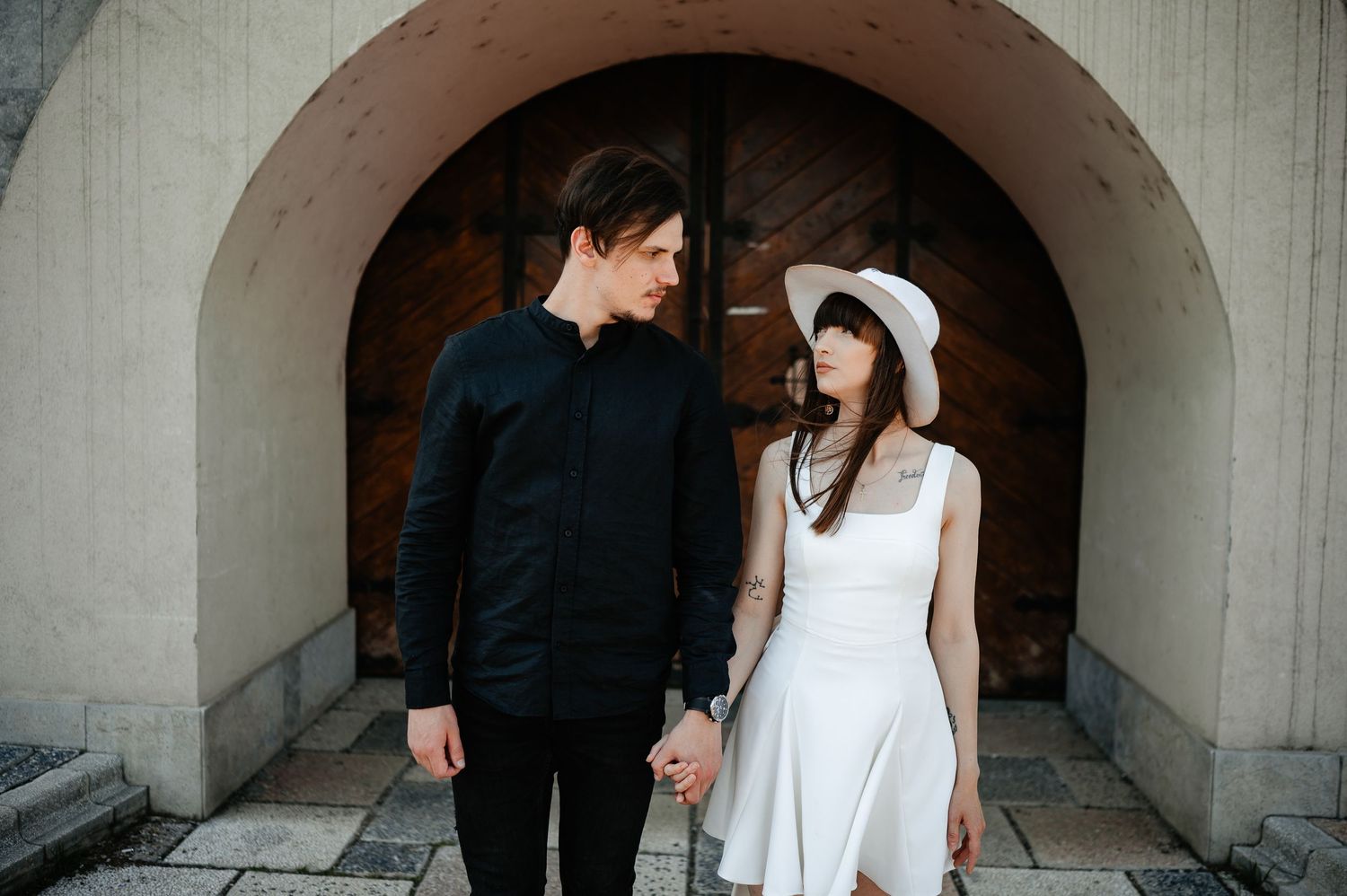 Young couple holding hands and gazing at each other under a stone archway with wooden doors.