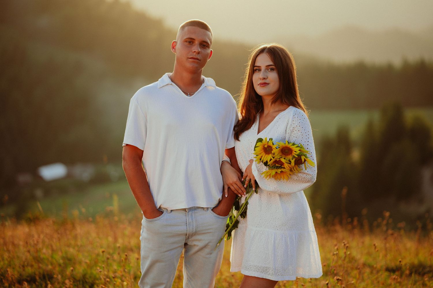 Young couple in white outfits standing together holding sunflowers in a golden meadow.