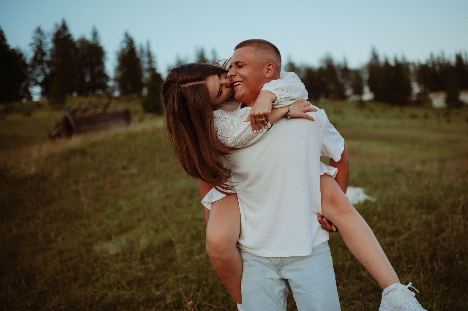 Happy couple embracing and laughing outdoors, man lifting woman in white dress on a green meadow with trees.