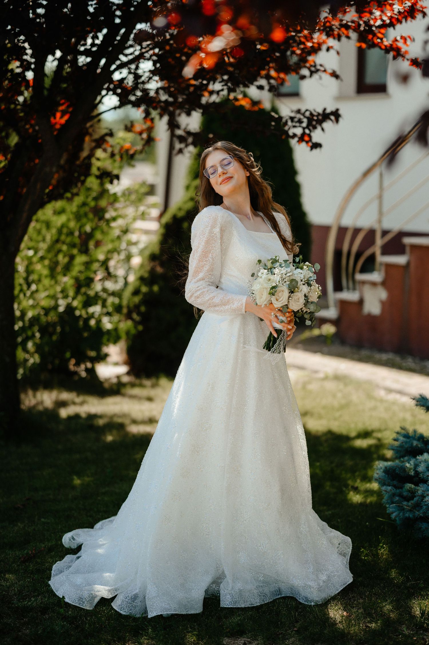 Beautiful bride in white lace wedding gown holding bouquet in sunlit garden setting.