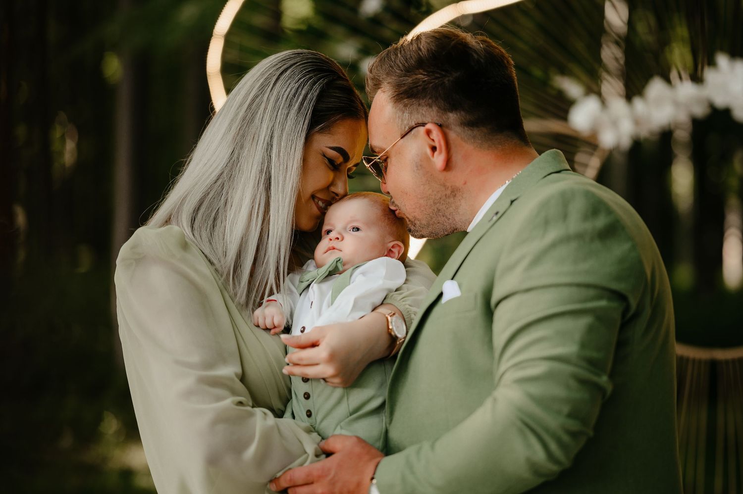 Parents in green attire tenderly kissing their newborn baby outdoors in a forest setting.