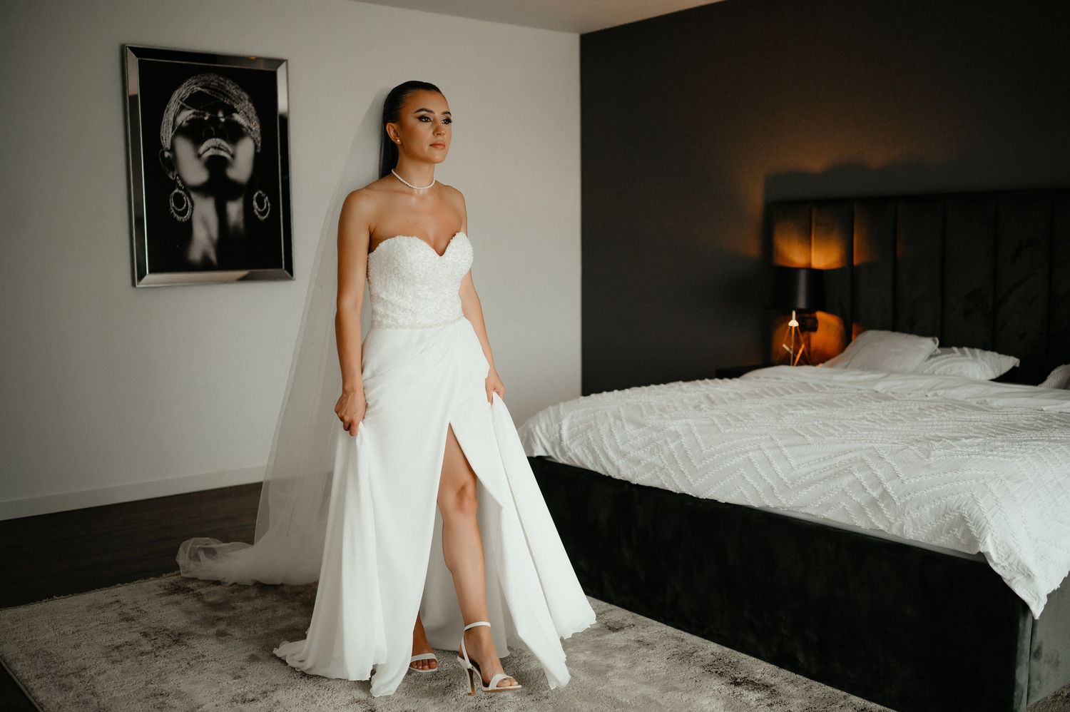 Bride in strapless white wedding gown with thigh-high slit poses elegantly in a modern bedroom.