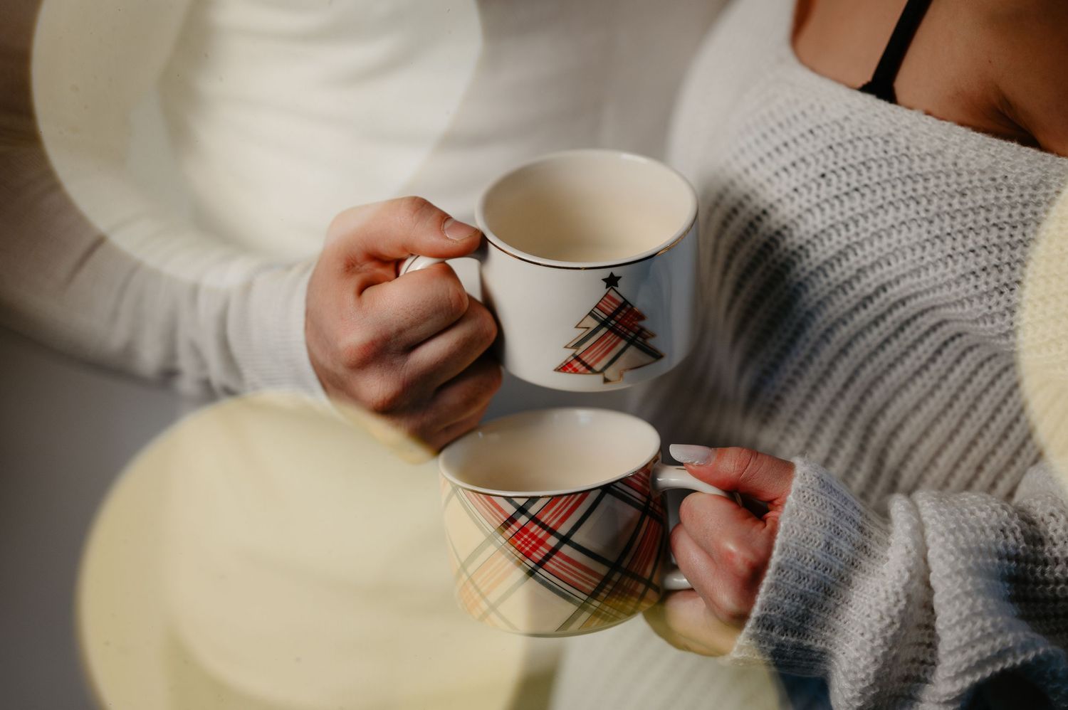 Two people holding festive plaid and Christmas tree mugs together during a cozy holiday gathering.