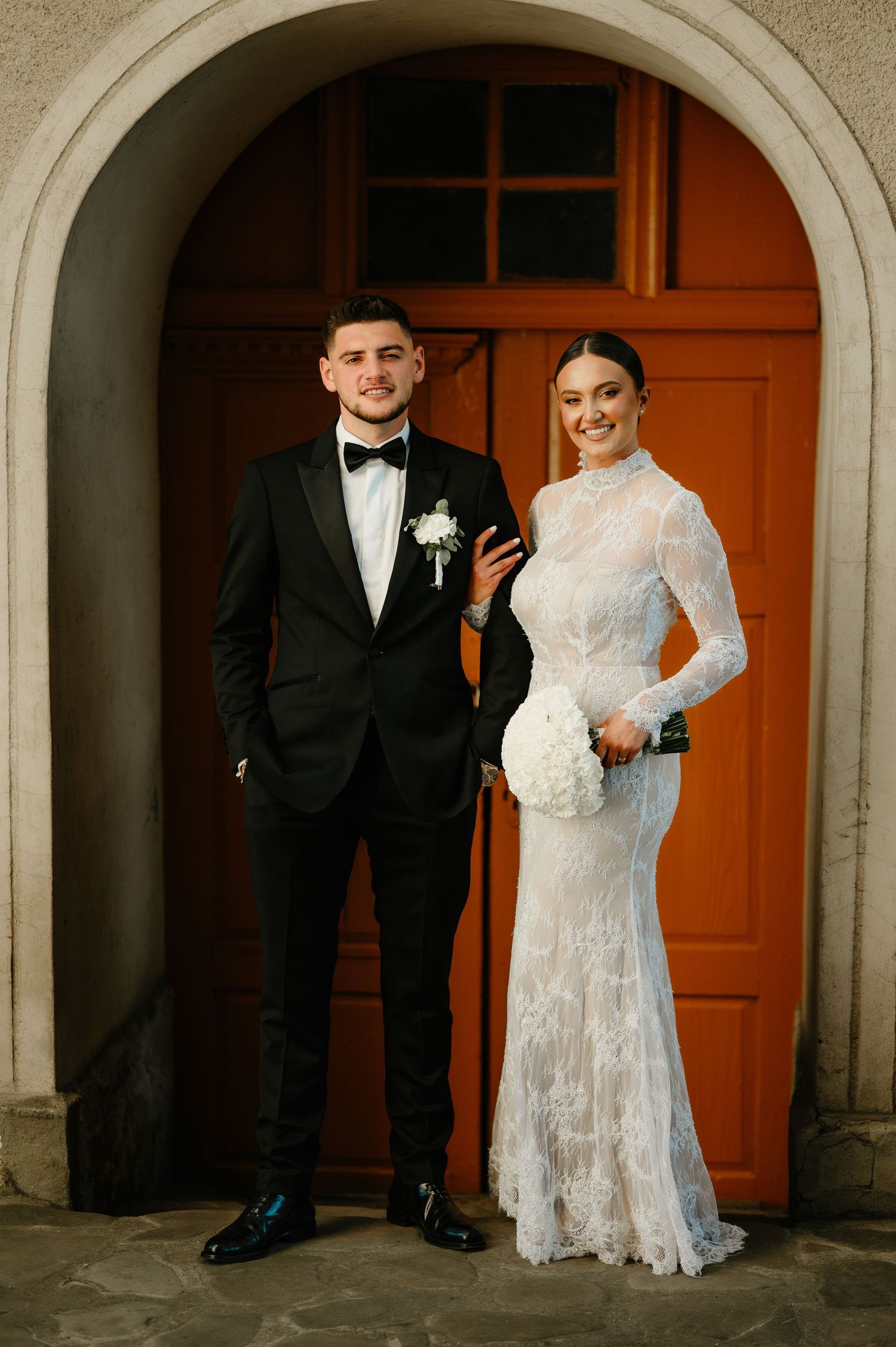 Elegant couple posing in front of an orange arched church door on their wedding day.