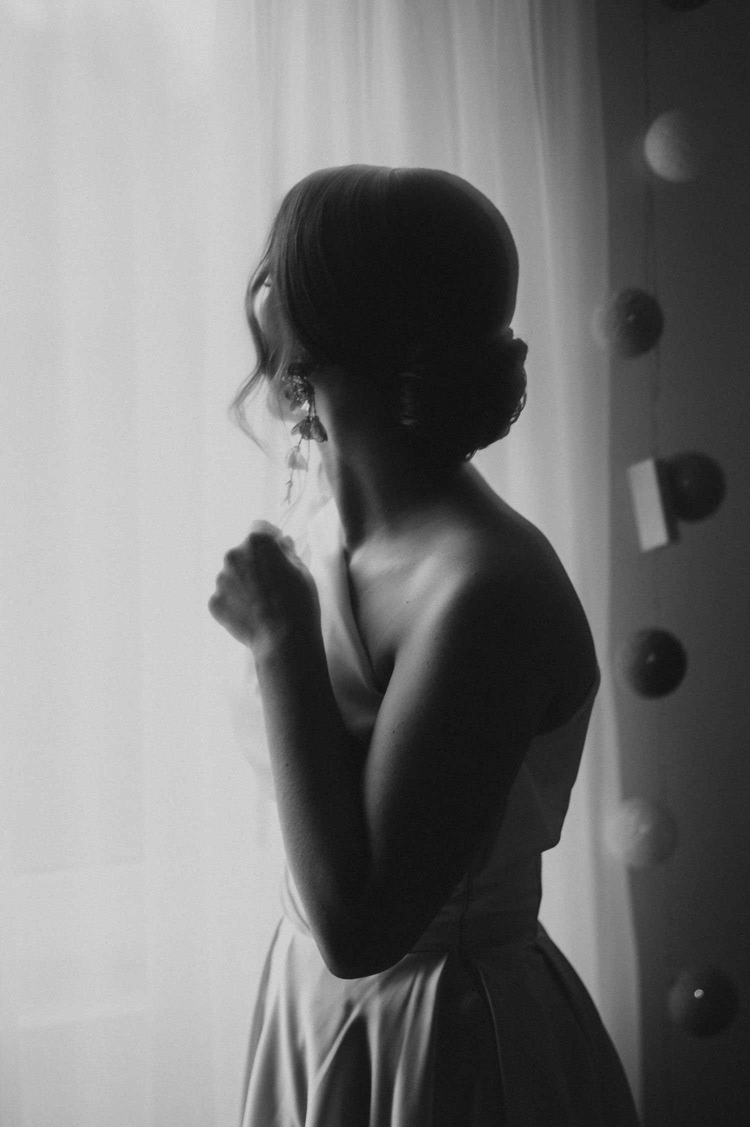 Elegant bride in strapless gown gazing out window in dreamy black and white wedding portrait.