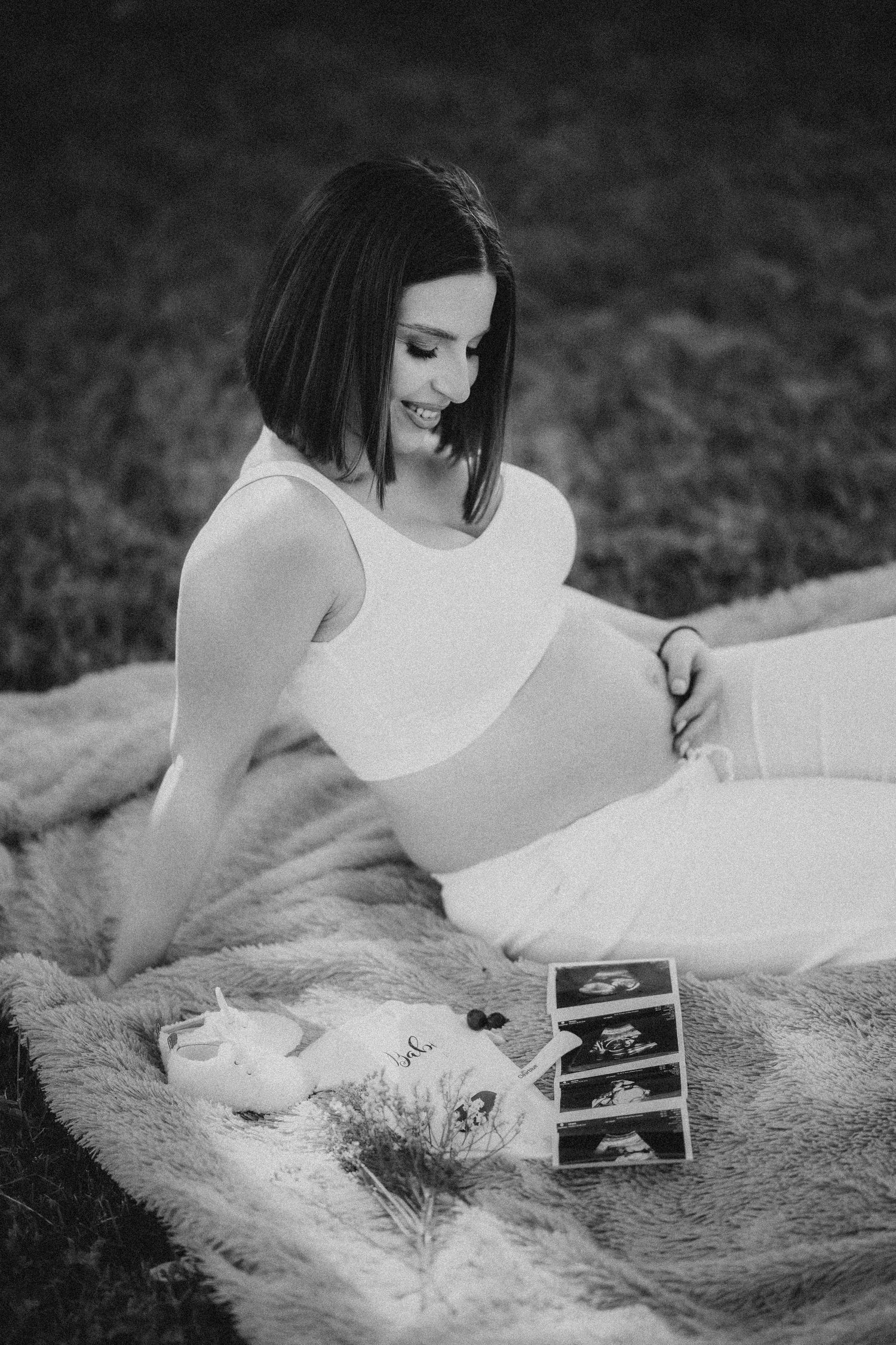 Pregnant woman in white outfit sits on blanket outdoors, smiling at ultrasound photos and baby shoes.