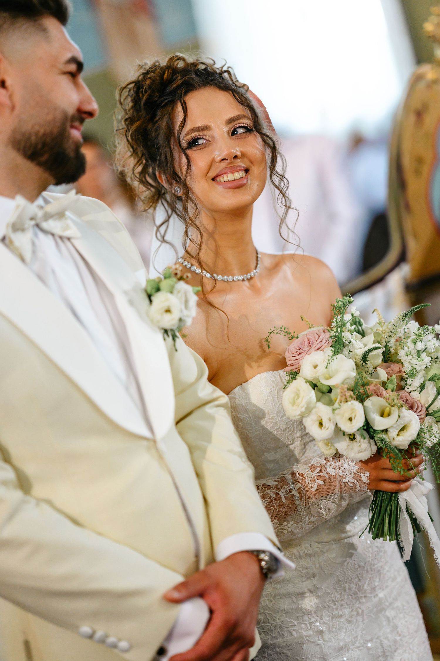 Smiling bride in strapless gown holds pink bouquet beside groom in ivory suit at wedding ceremony.