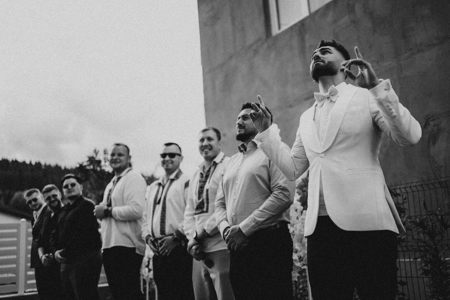 Black and white photo of groomsmen in white suits laughing and celebrating outdoors before a wedding ceremony.