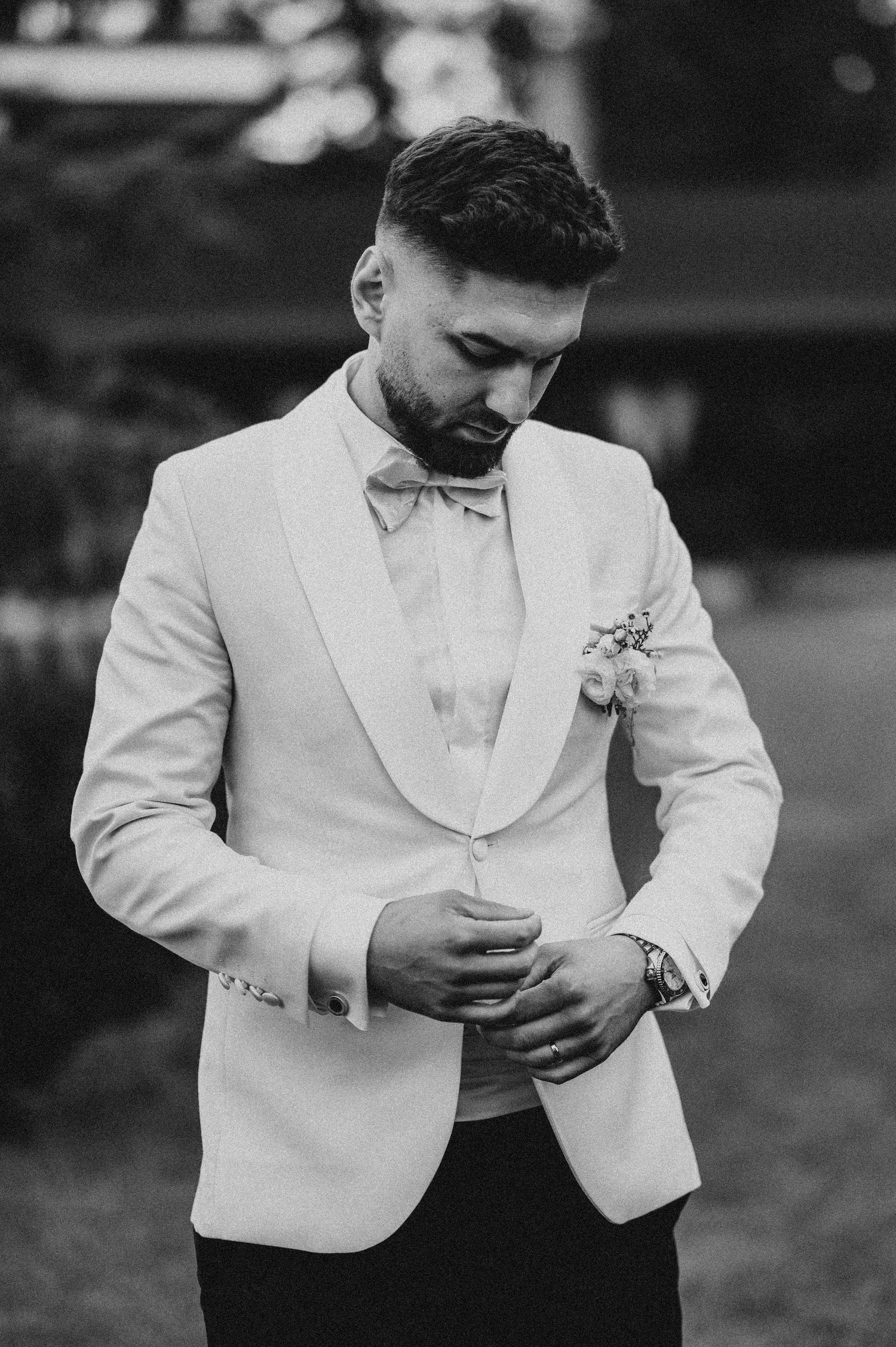 A groom in a white tuxedo jacket and bow tie adjusts his button while wearing a boutonniere outdoors.