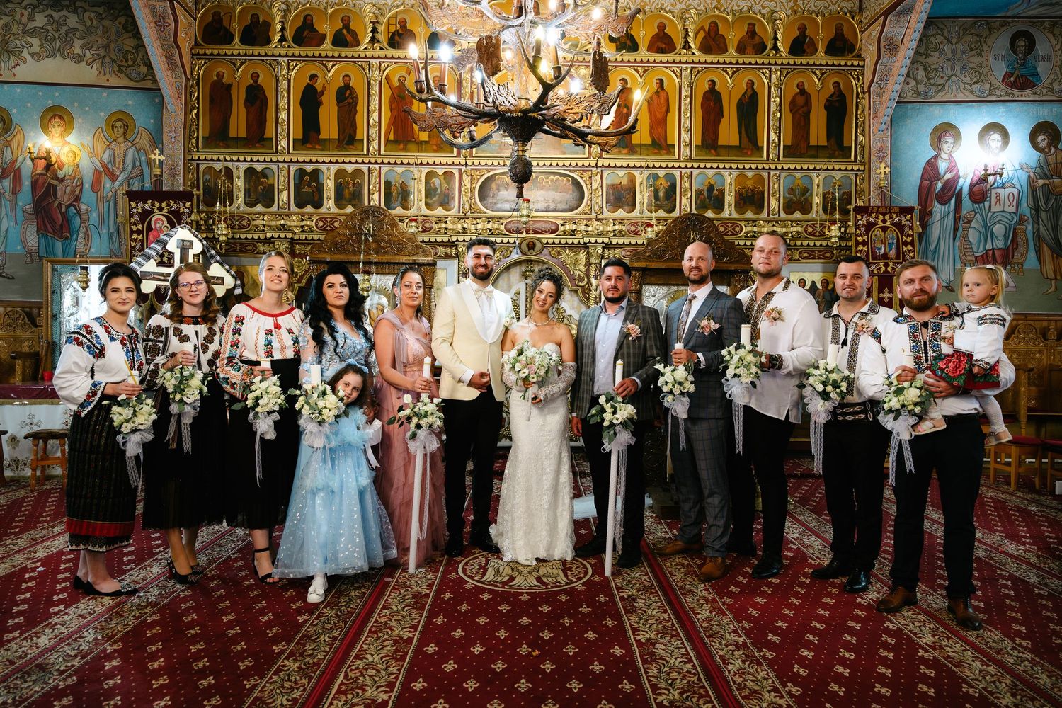 Wedding party poses in ornate Orthodox church, bride and groom flanked by bridesmaids and groomsmen in traditional Romanian attire.