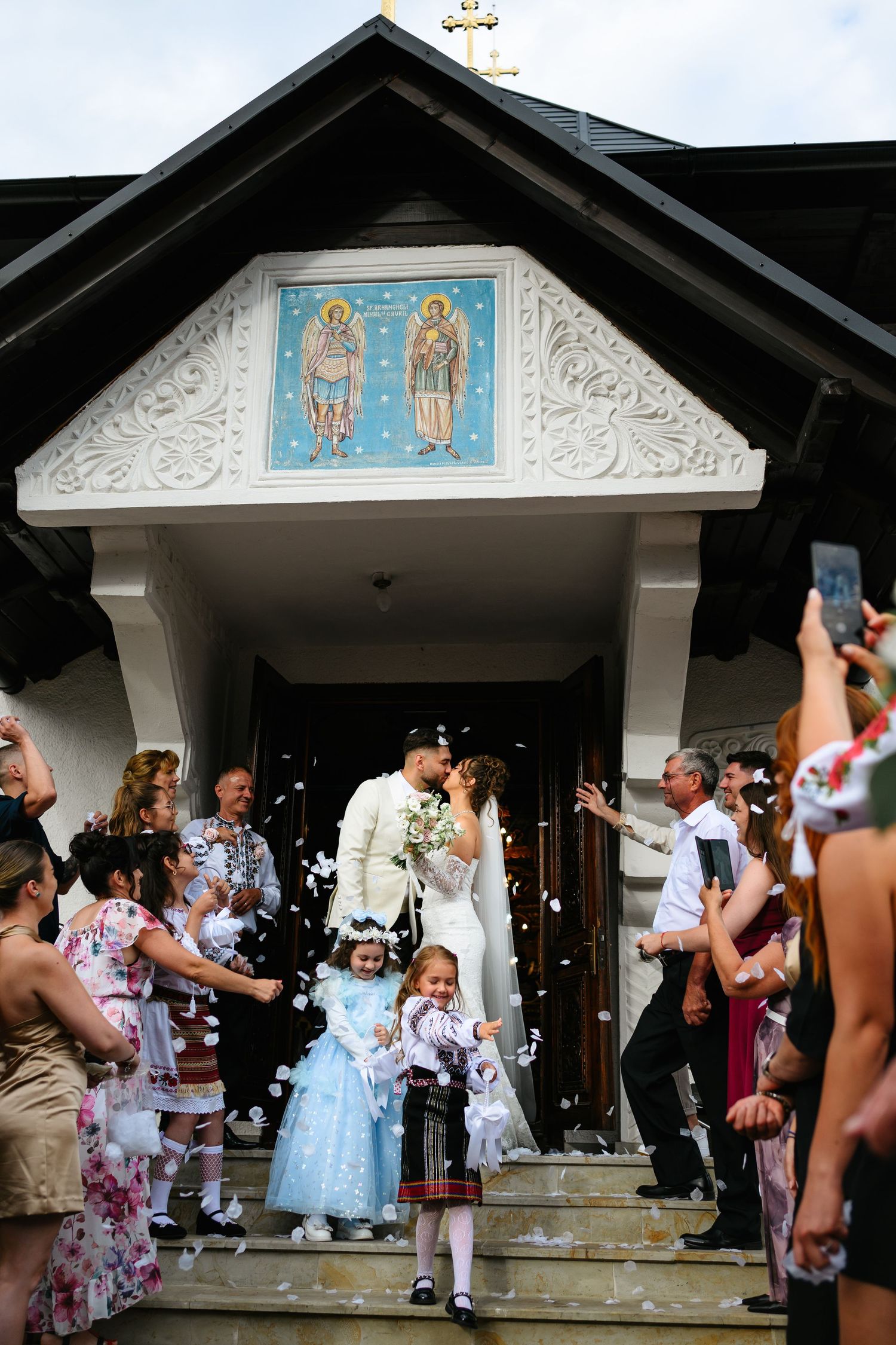 Bride and groom kissing on church steps while guests throw confetti at their wedding ceremony.