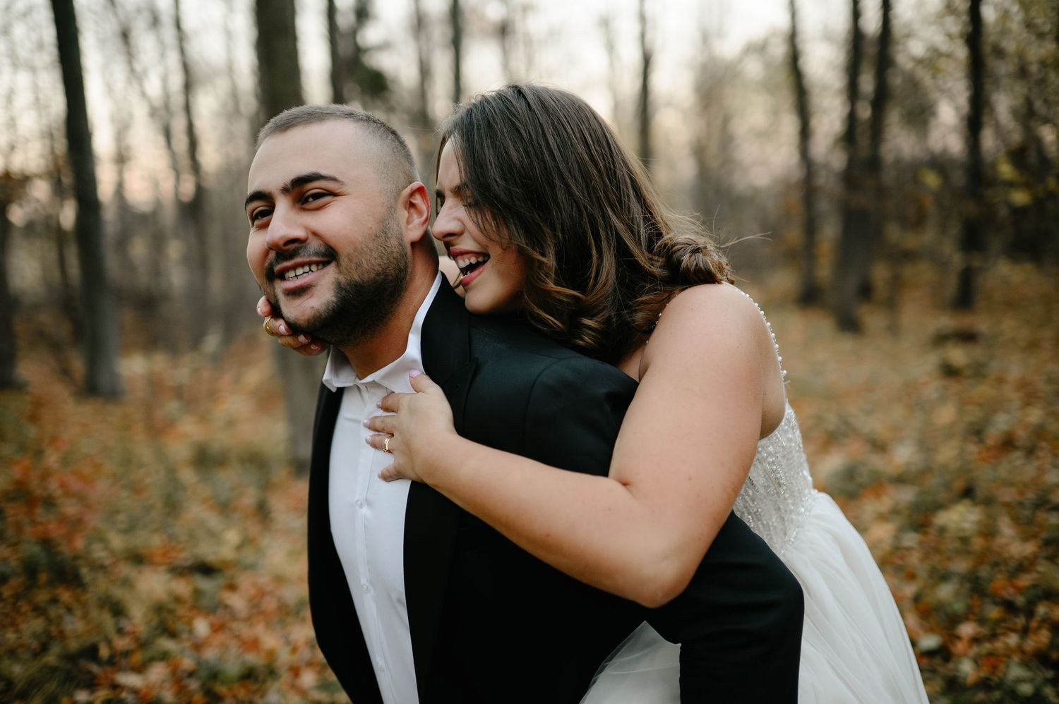 A joyful bride in a white gown laughs while riding piggyback on a groom in a black tuxedo outdoors.