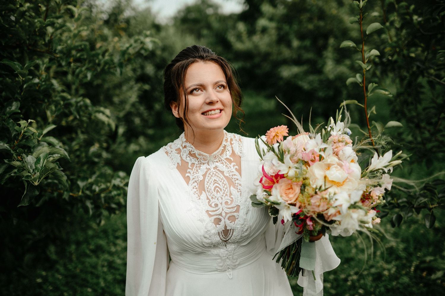 Smiling bride in lace high-neck wedding gown holds vibrant peach and pink bouquet among green orchard trees.