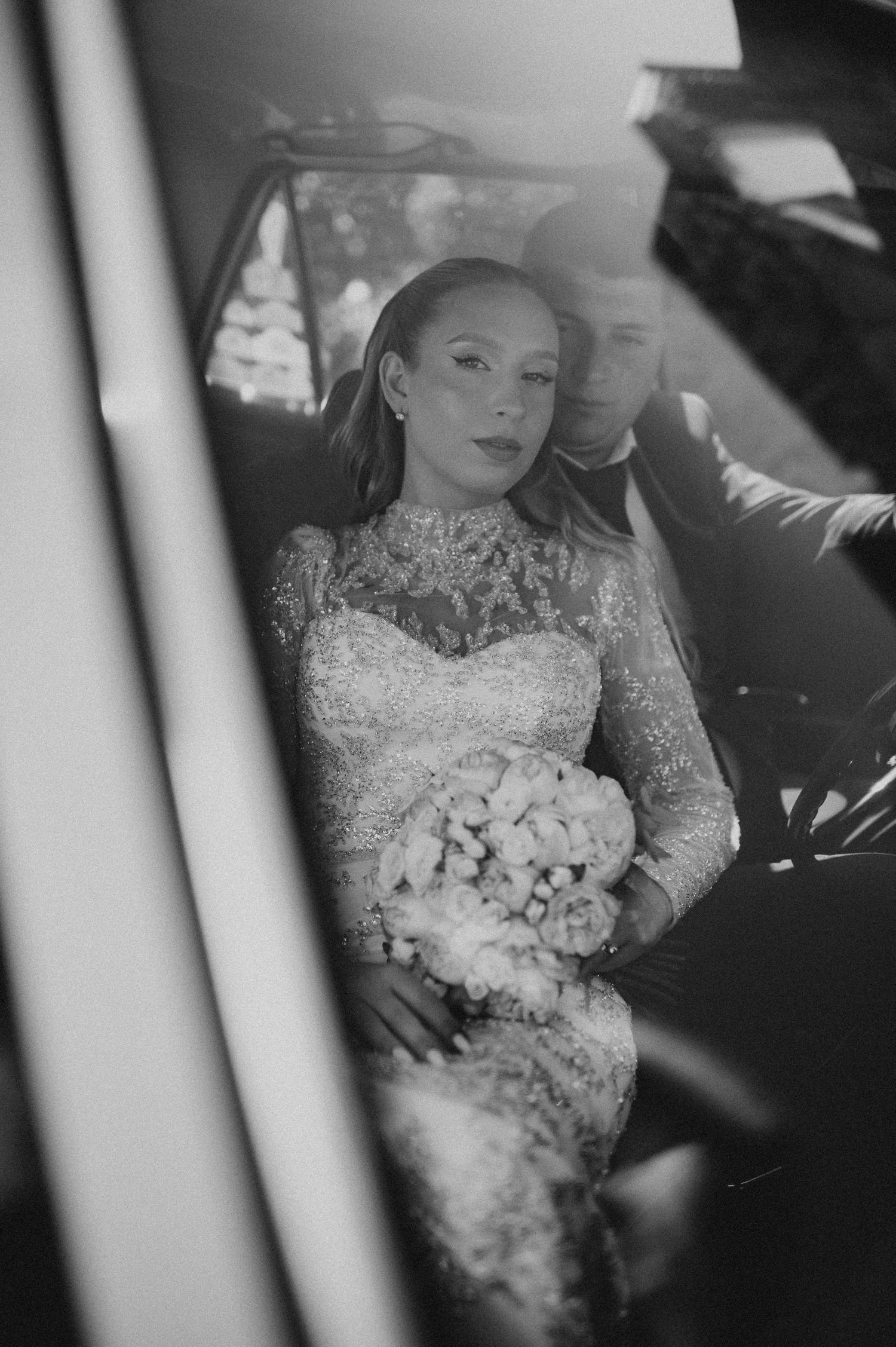 Black and white photo of a bride in lace gown holding bouquet sitting with groom in a wedding car.
