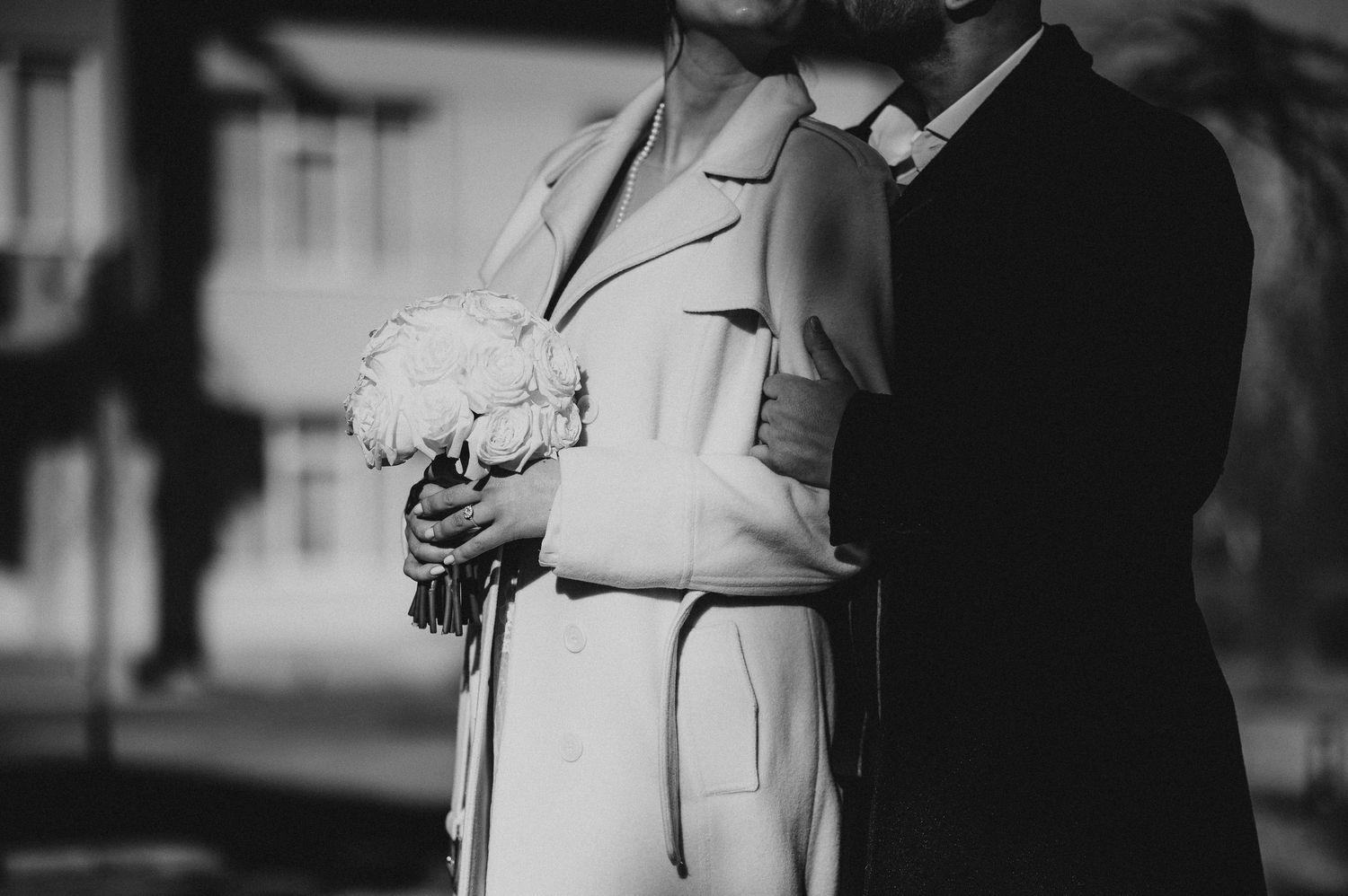 Black and white photo of a couple embracing, bride holding a white bouquet in an elegant coat outdoors.