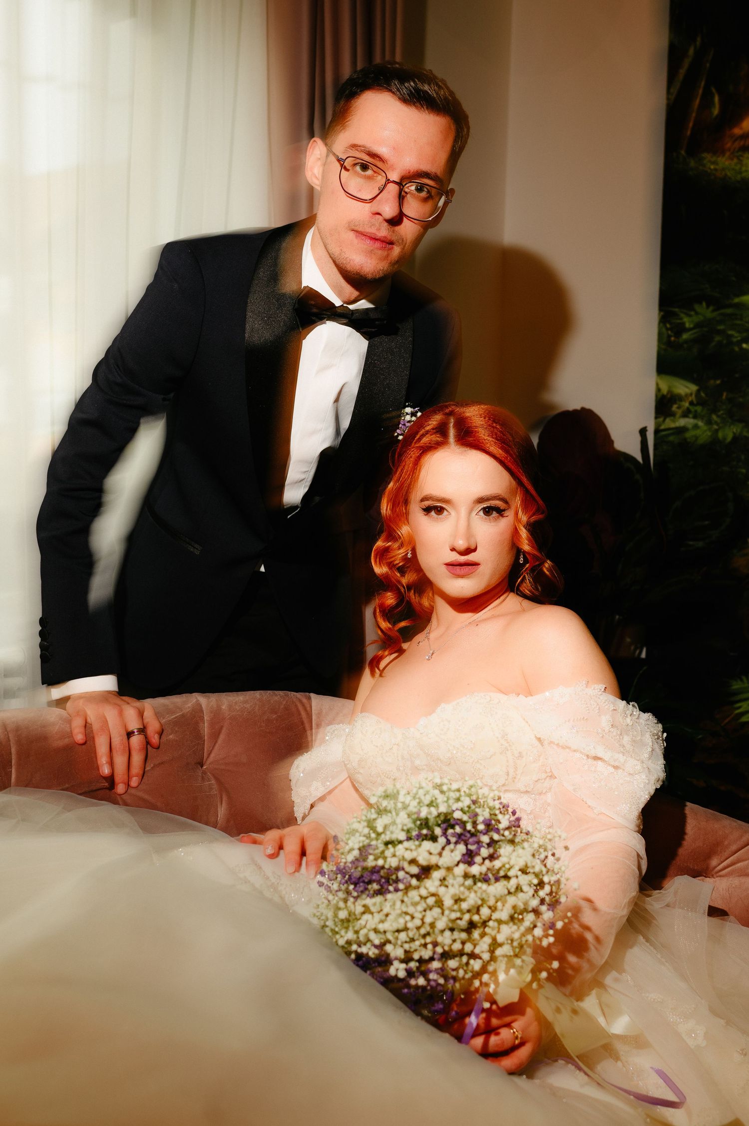 A groom in a black tuxedo stands behind a red-haired bride in a white off-shoulder gown holding a floral bouquet.