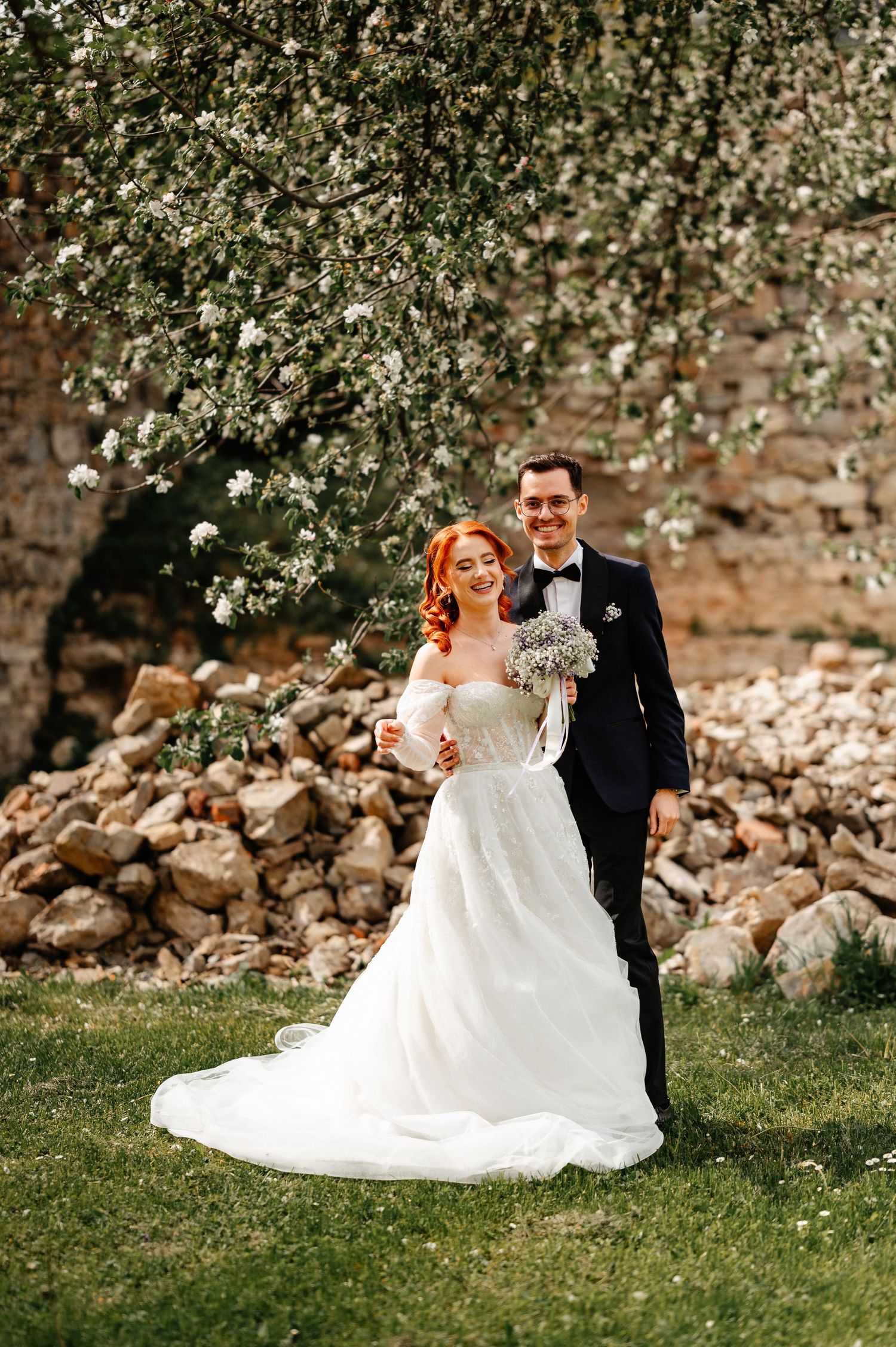 A smiling bride in a white gown and groom in navy suit pose together under blooming trees near a stone wall.