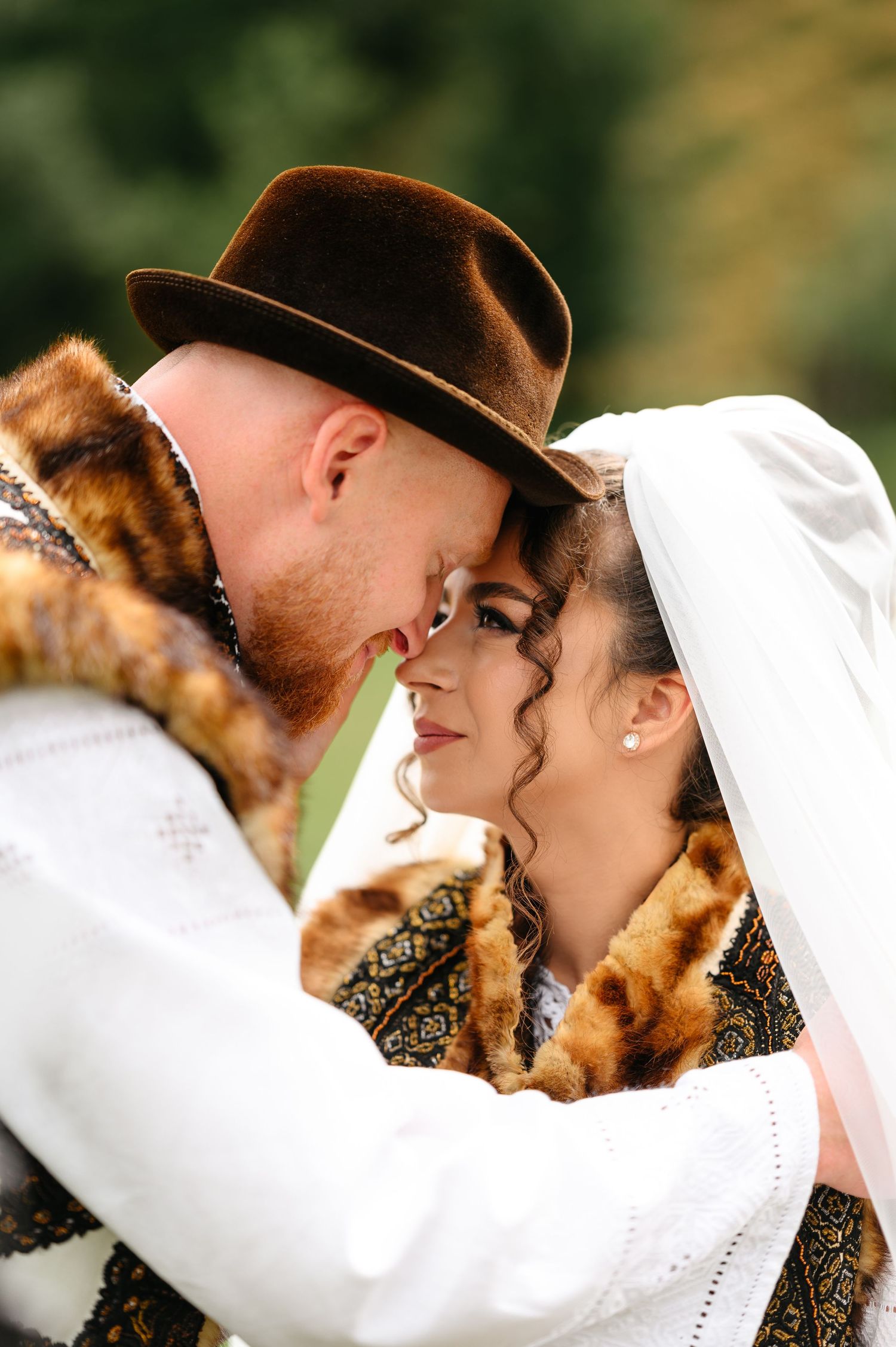 A couple in traditional Eastern European wedding attire share a romantic moment, the groom in a fur coat and brown hat.