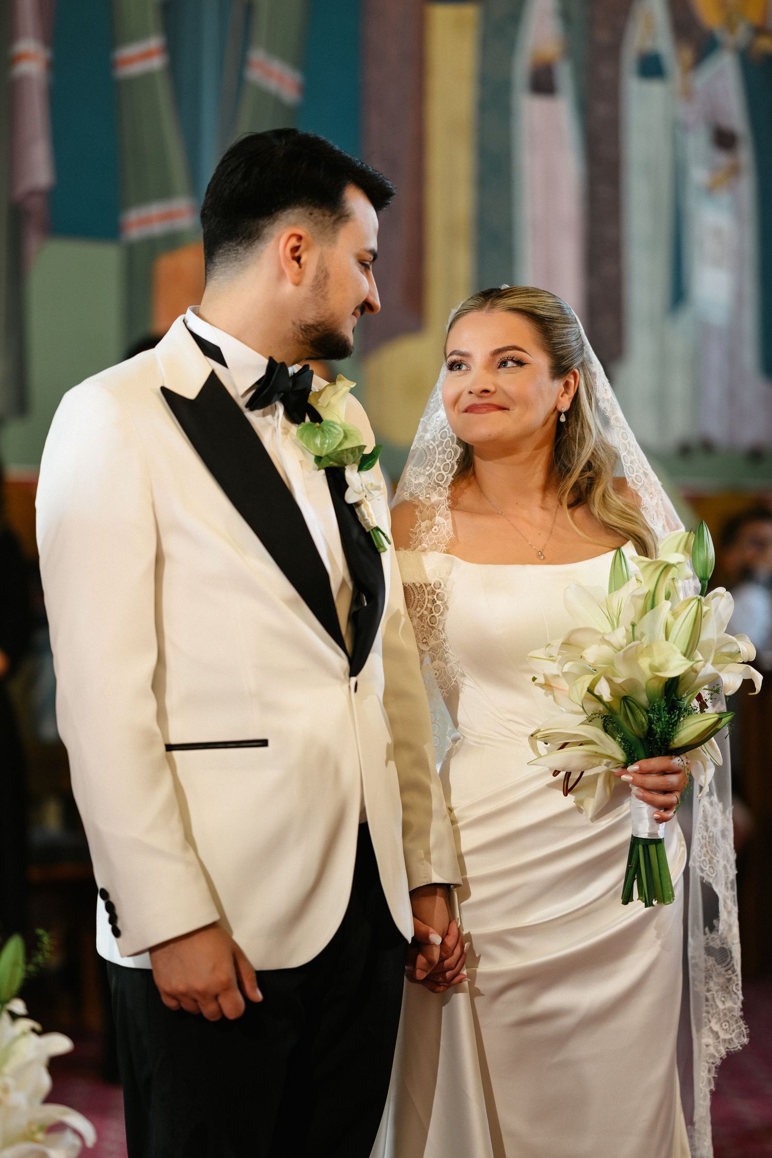 Bride and groom holding hands at church altar, bride in white gown with veil holding lily bouquet, groom in white tuxedo.