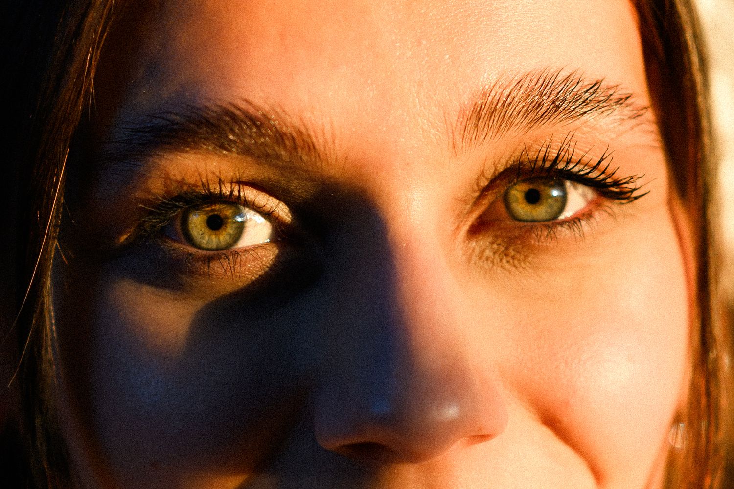Close-up portrait of a young woman with striking green eyes and dark eyebrows in dramatic lighting.