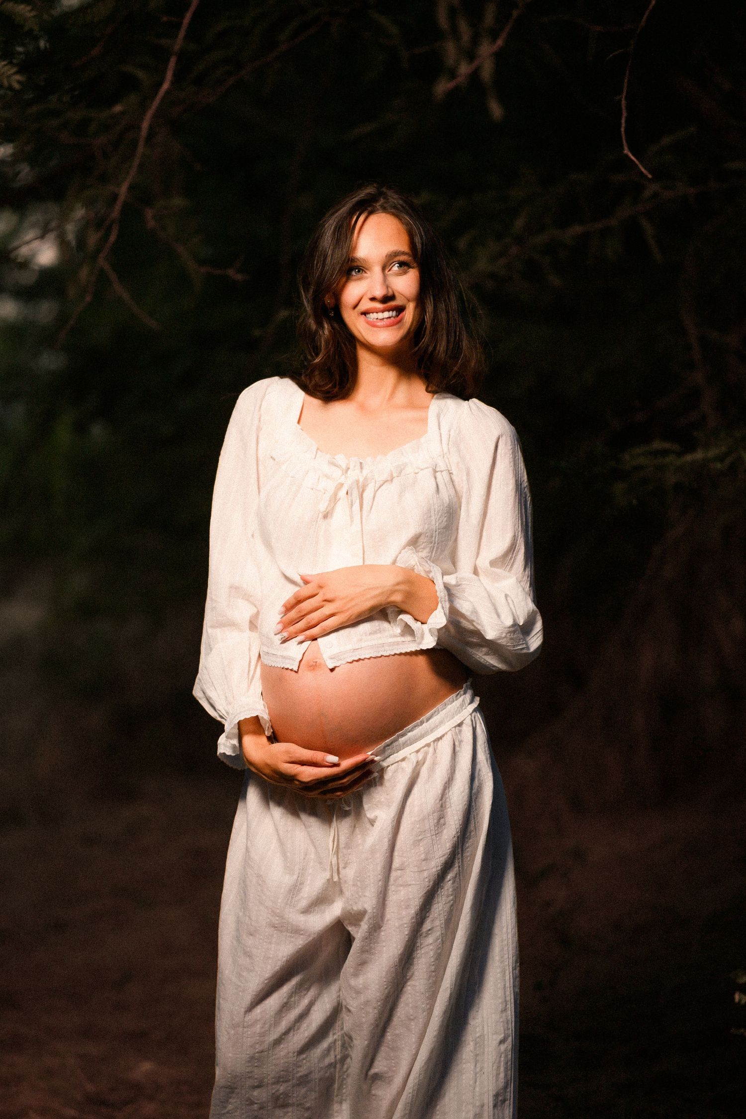Smiling pregnant woman in white dress cradling her baby bump outdoors in a sunlit forest setting.