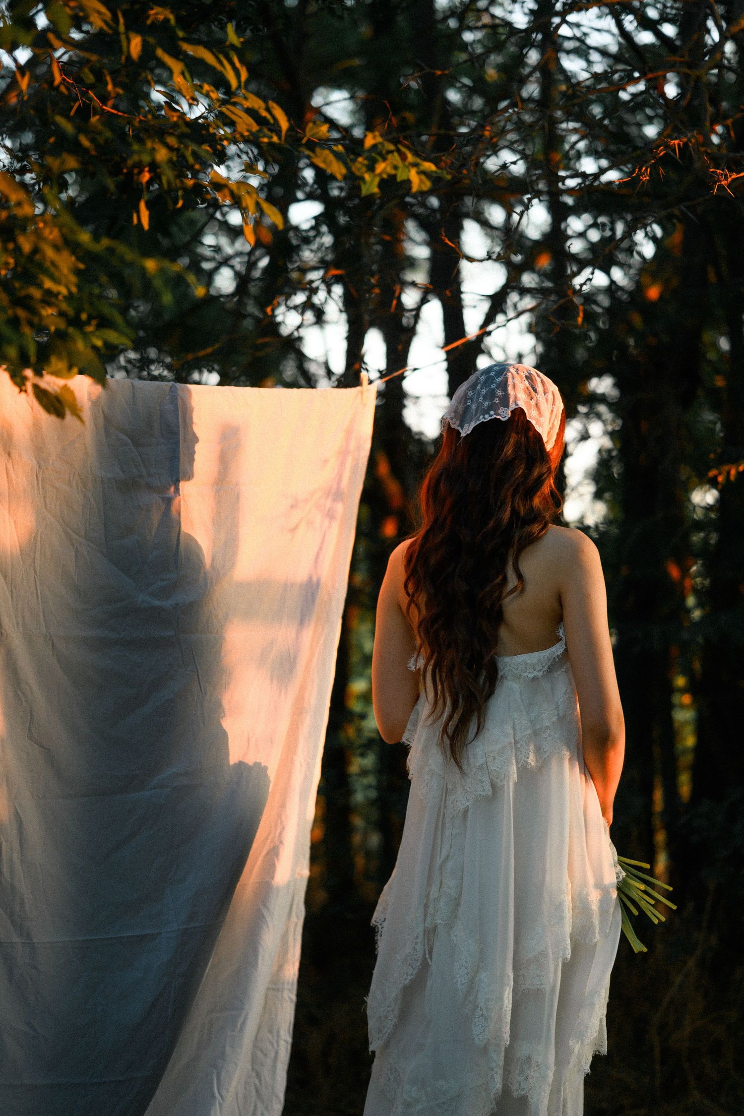 Woman in white dress and lace headband stands near hanging white fabric in a sunlit forest at golden hour.
