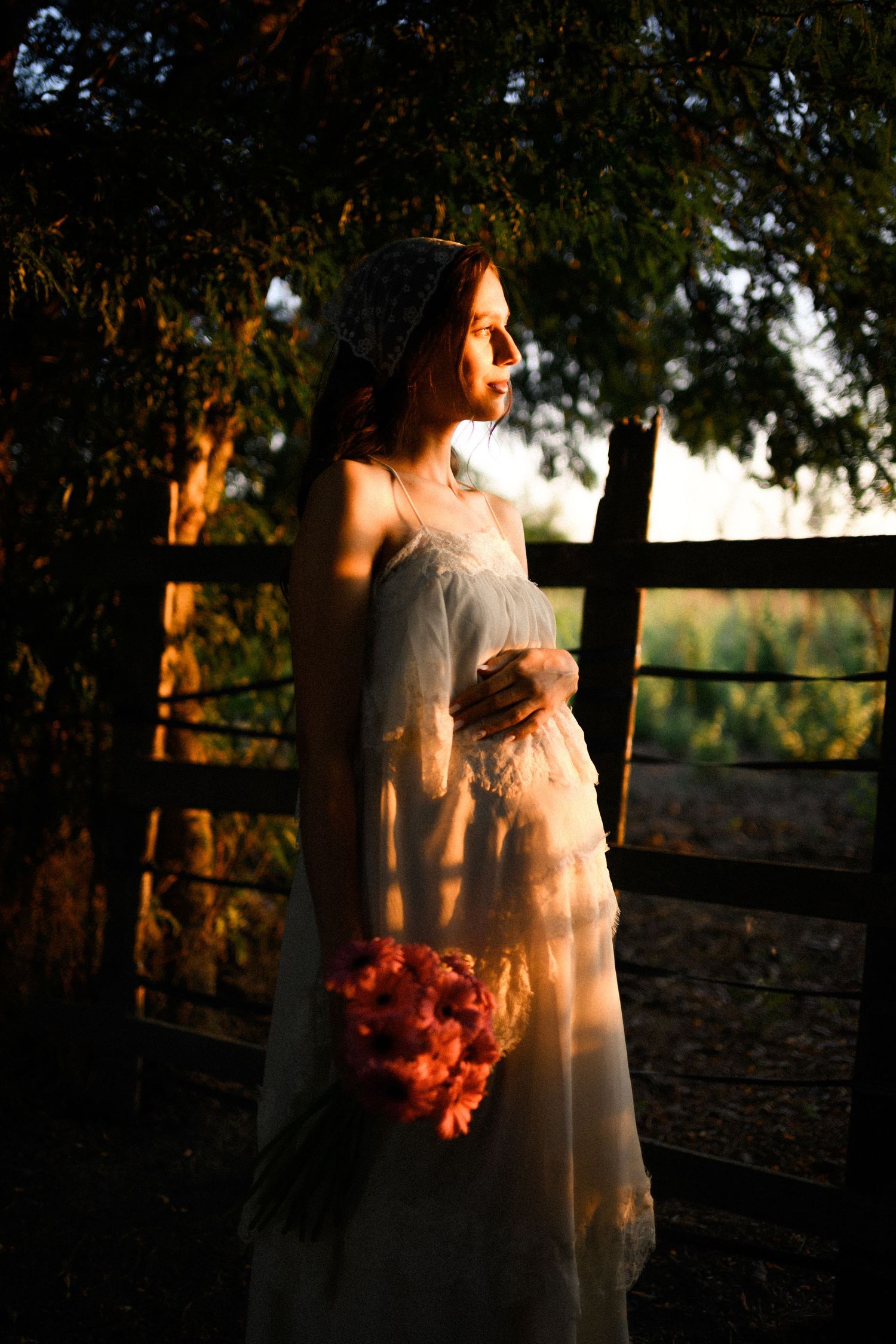 Pregnant bride in white lace gown holds pink bouquet at golden hour by rustic wooden fence surrounded by trees.