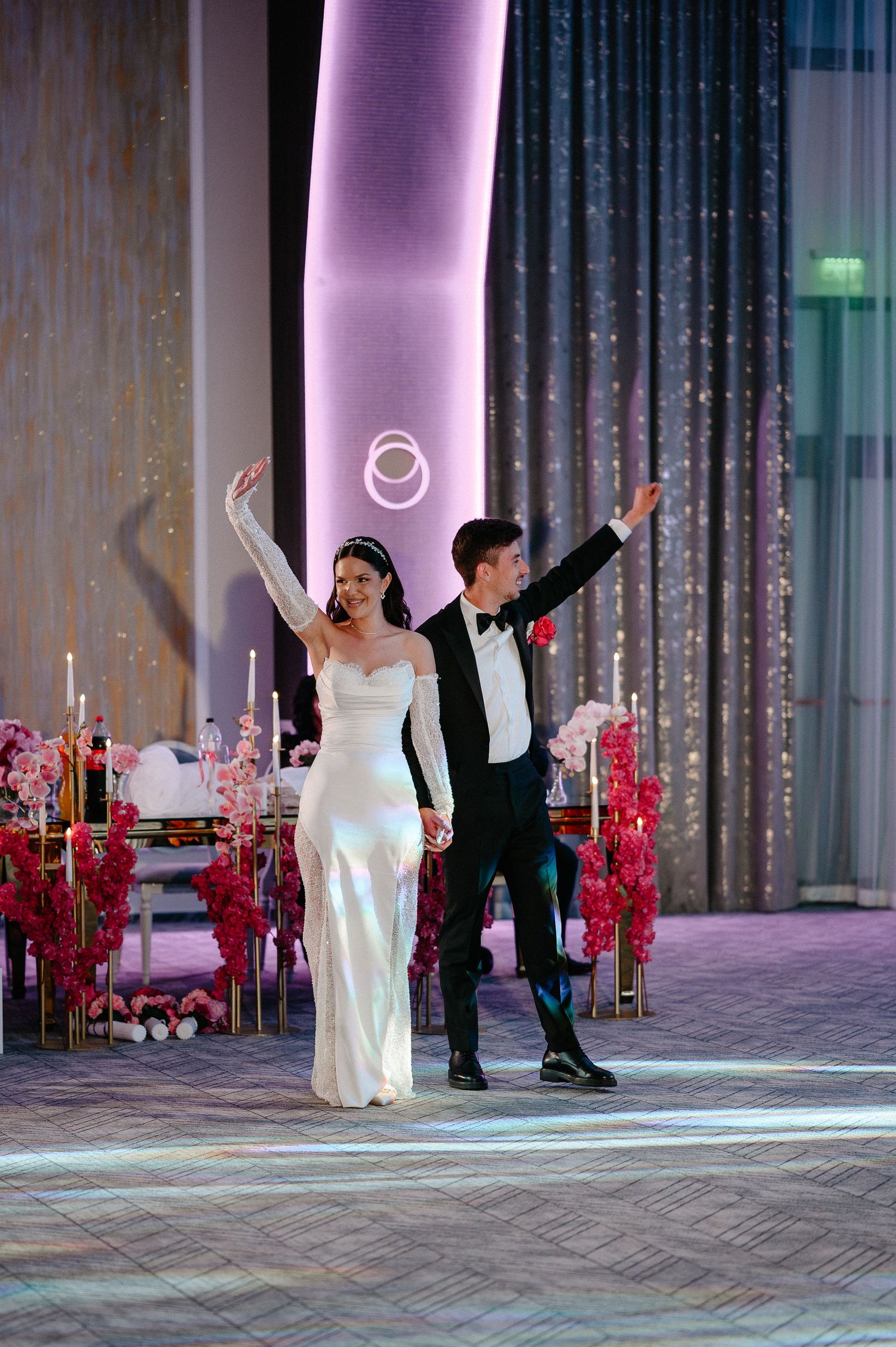 Bride in white gown and groom in tuxedo make grand entrance at elegant wedding reception with pink decor.