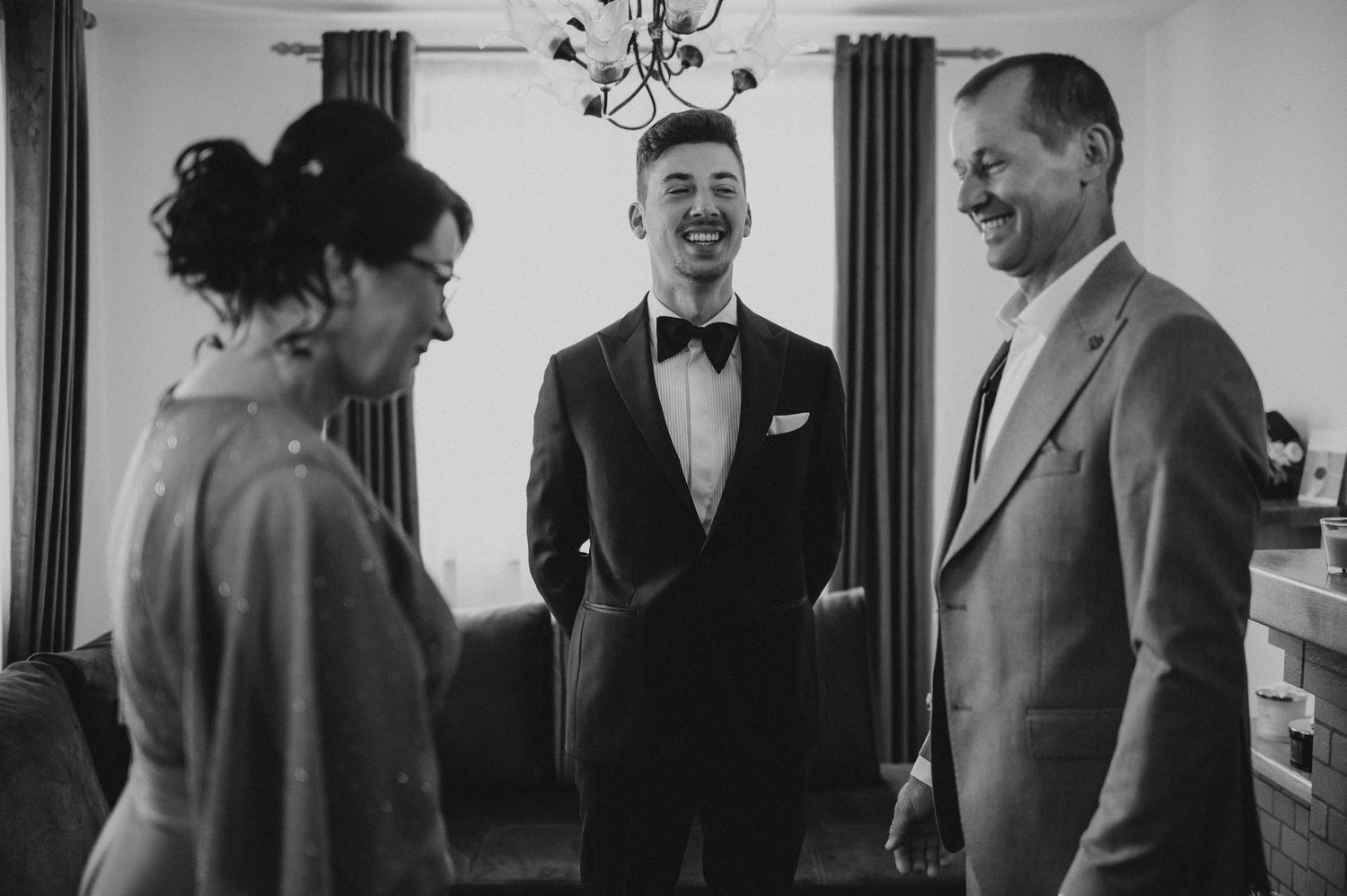 Groom in tuxedo laughing with parents in black and white wedding preparation photo.