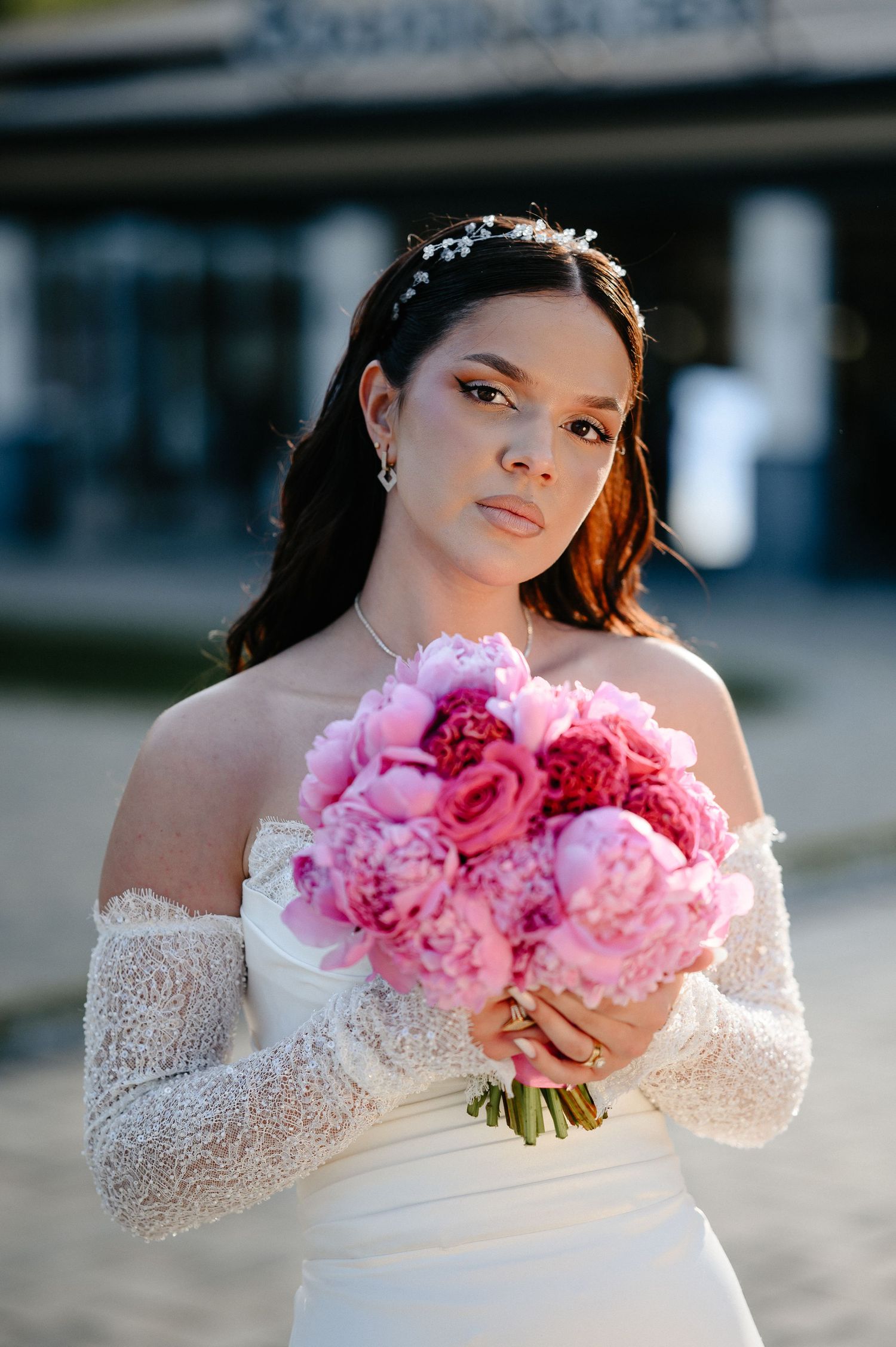 Close-up portrait of bride in white lace gown holding lush pink peony bouquet with pearl hair accessory.