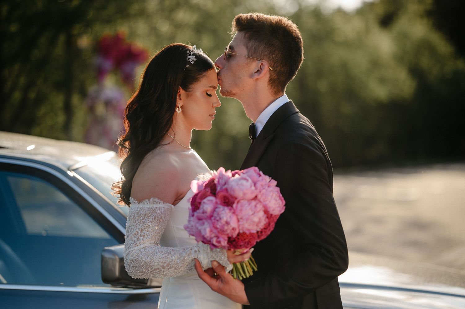 Bride and groom share tender moment face-to-face, bride holding vibrant pink peony bouquet outdoors.