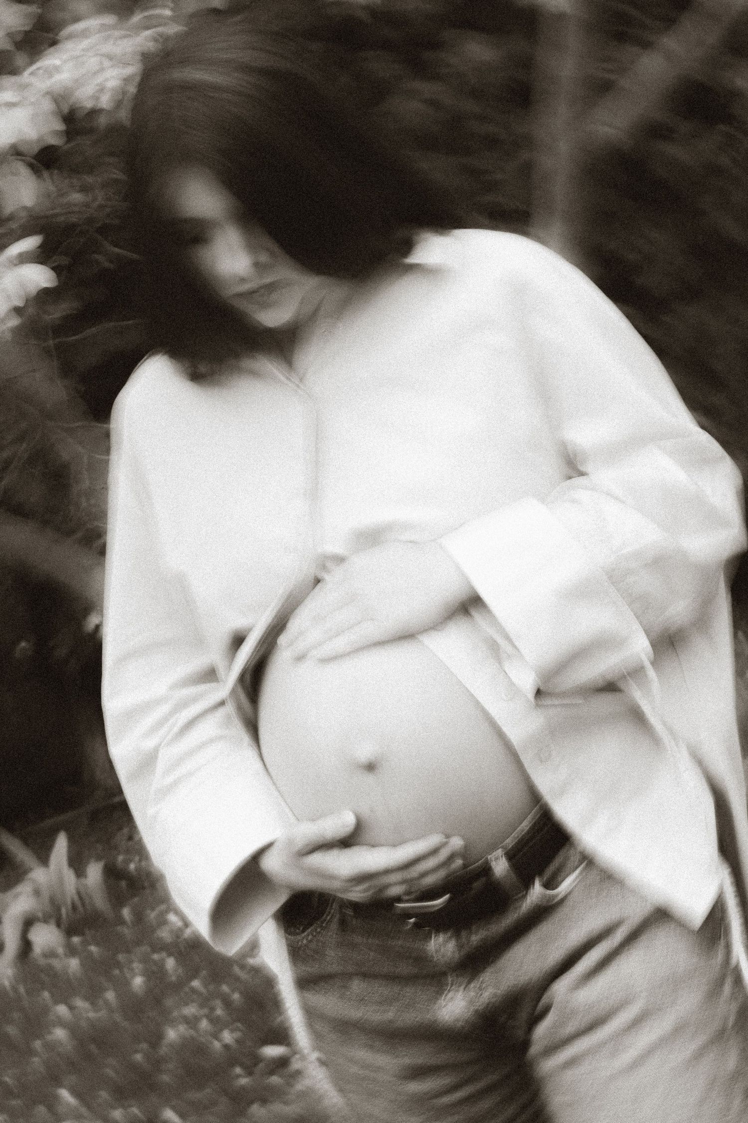 Black and white photo of pregnant woman in white shirt cradling baby bump outdoors.