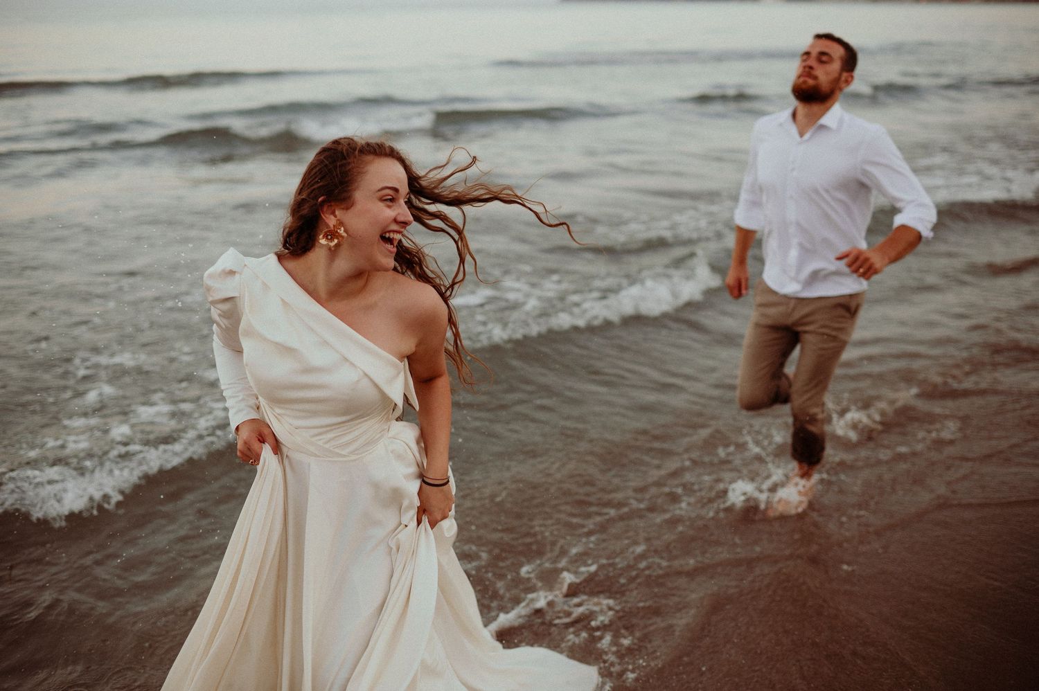 Bride running on beach with groom chasing her through ocean waves at sunset.
