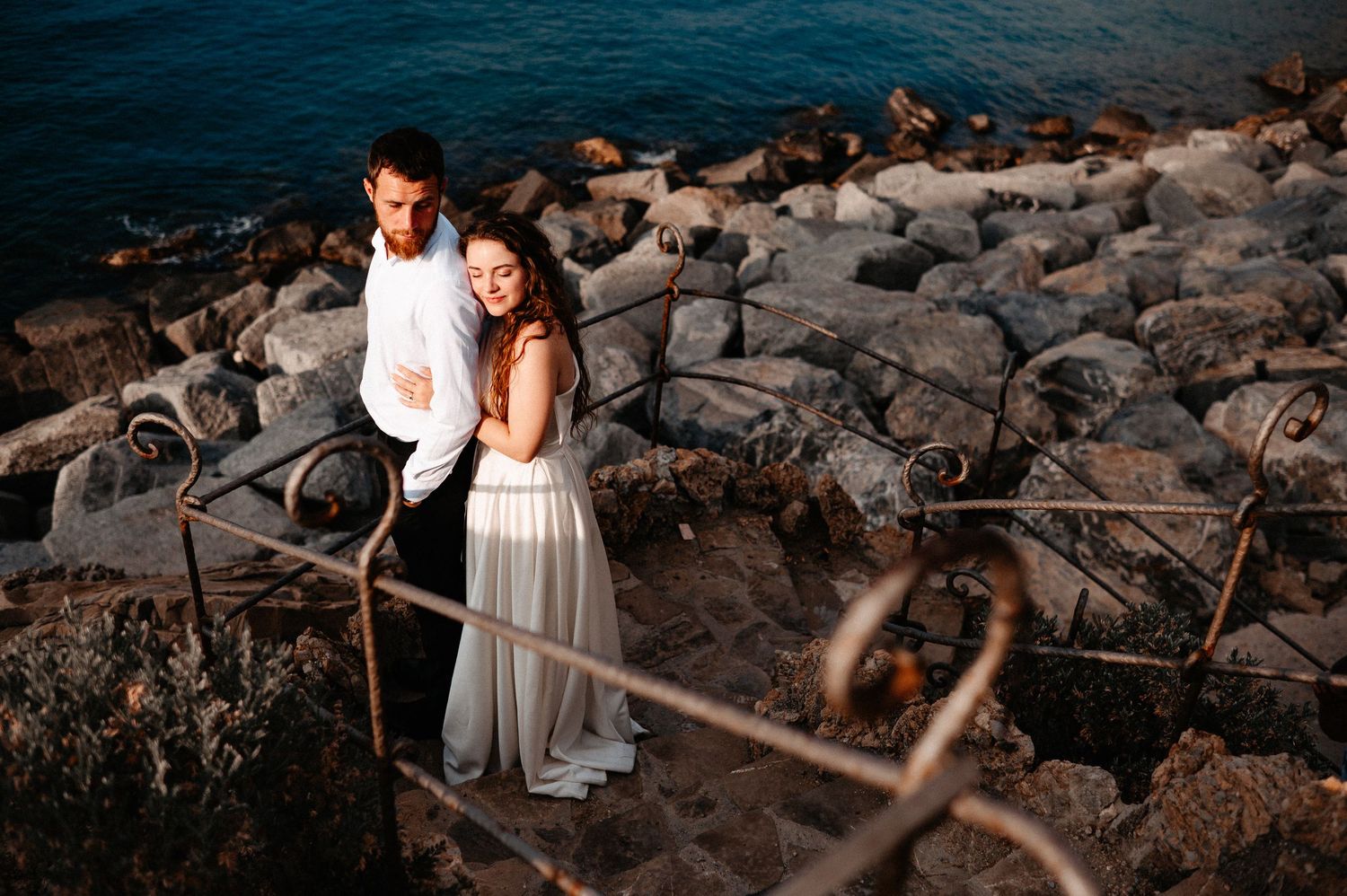 Wedding couple embracing on rocky Mediterranean coastal stairway at golden sunset hour.