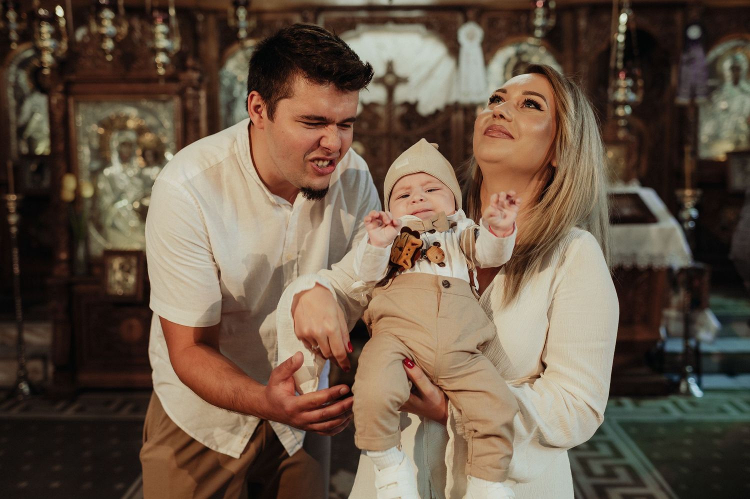 Young couple holds smiling baby in beautifully decorated Orthodox church setting.