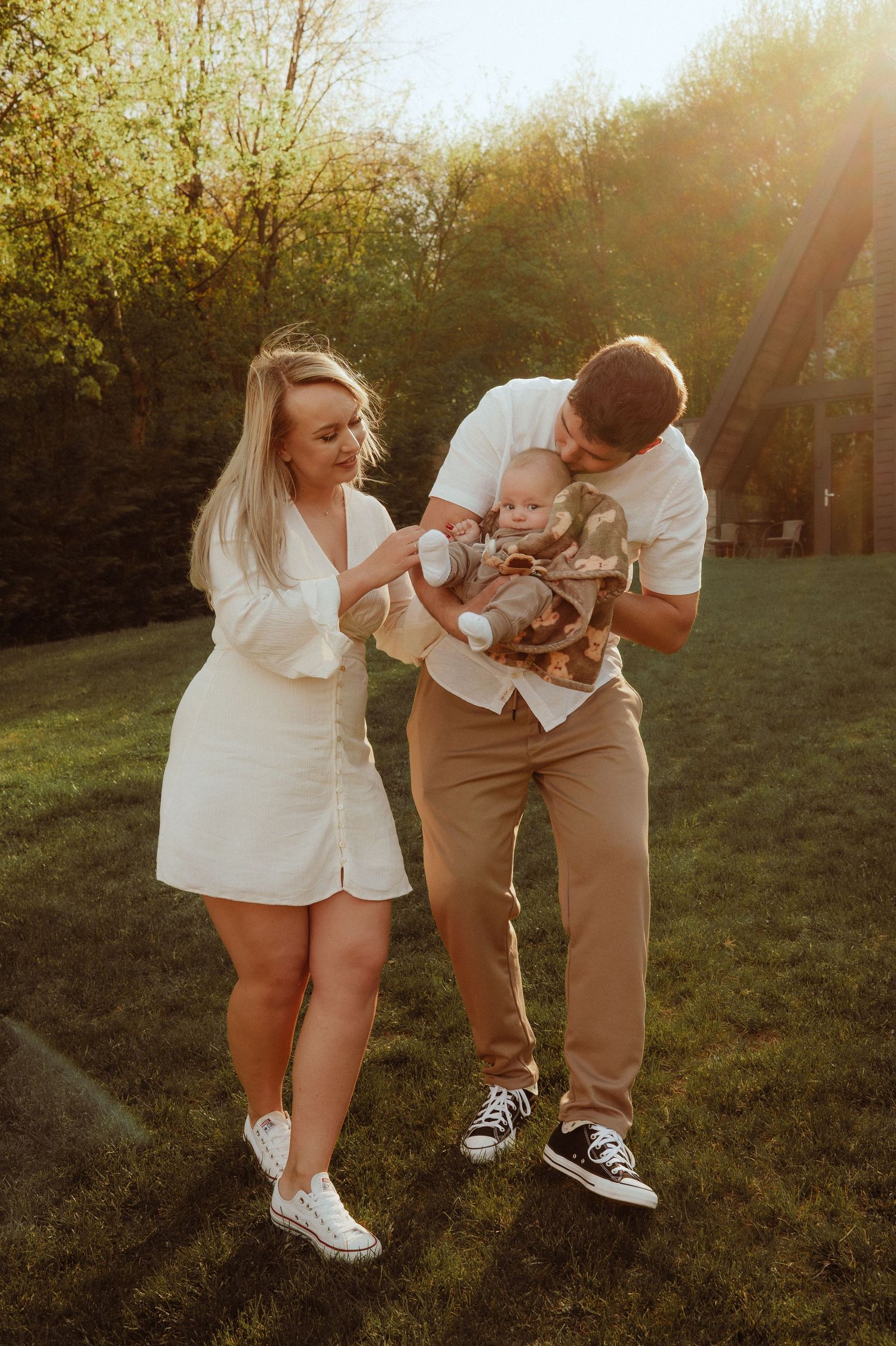 Young couple in white and beige outfits lovingly holding their newborn baby in a sunny garden.