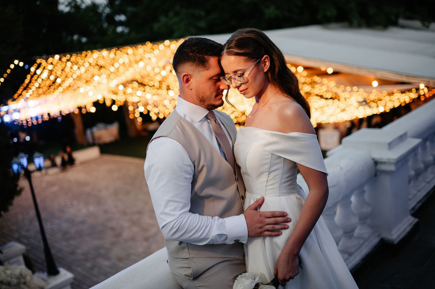 Bride and groom embracing tenderly under a canopy of warm fairy lights at their outdoor wedding.