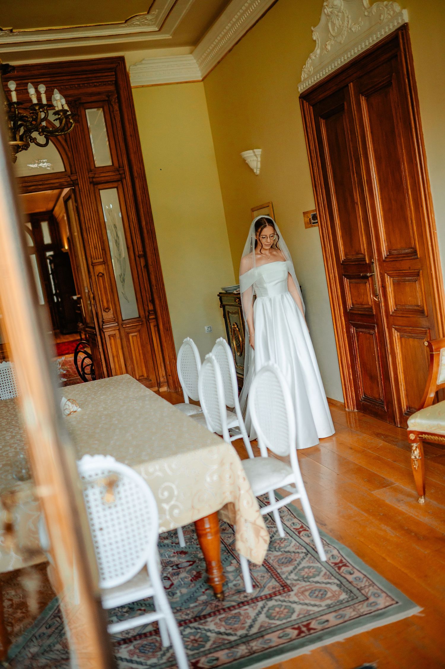Bride in off-shoulder white wedding dress and veil stands in ornate vintage dining room.