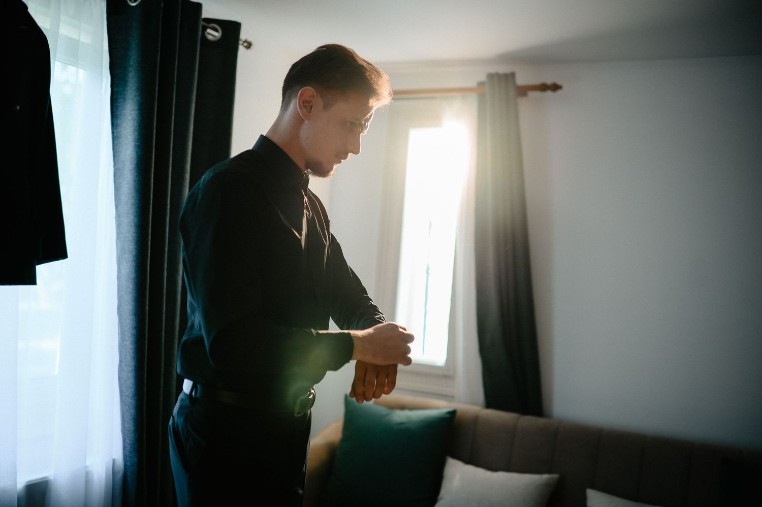 Groom in black attire checking his watch by a bright sunlit window in a room.