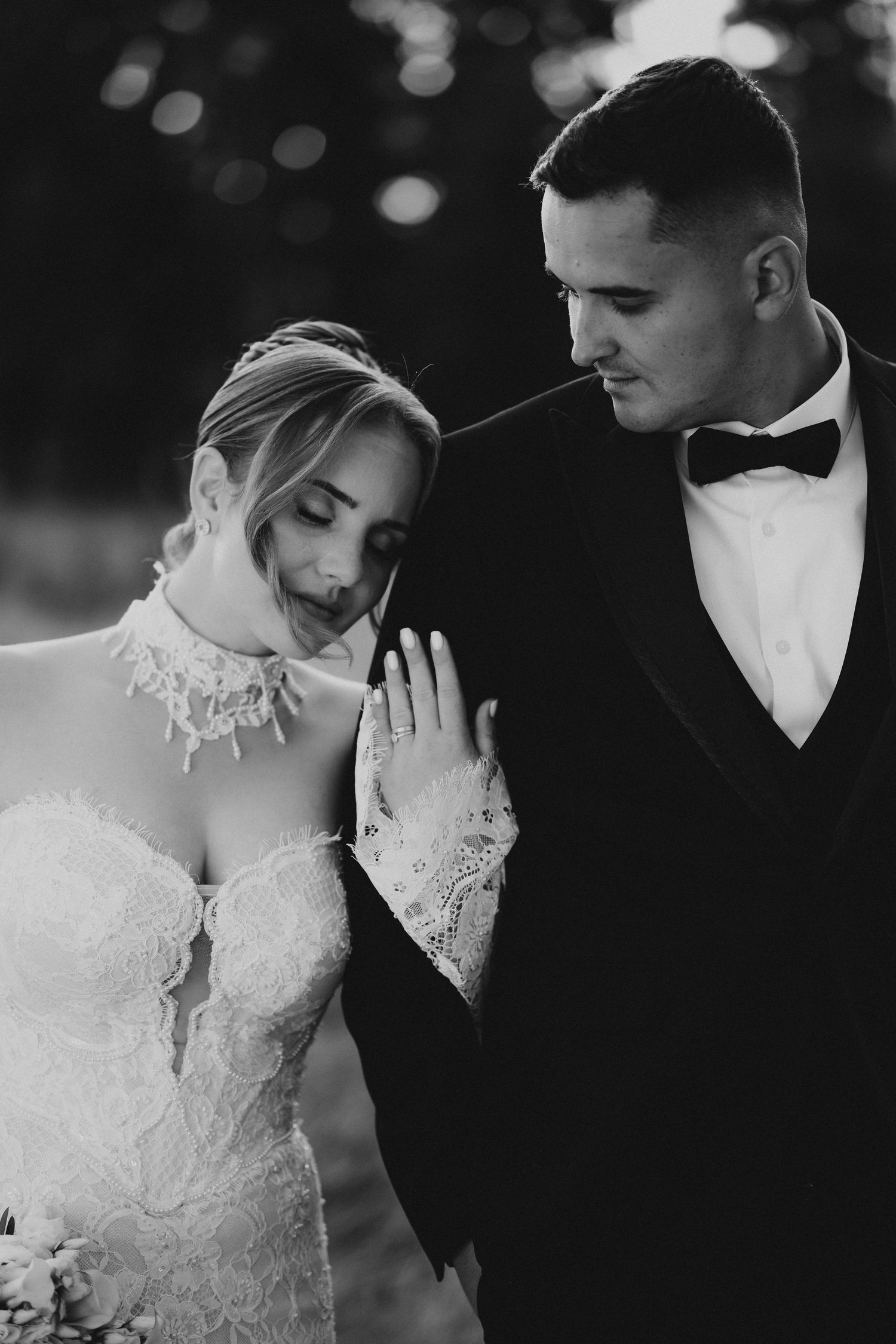 Black and white wedding portrait of bride in lace gown and groom in tuxedo sharing an intimate moment outdoors.