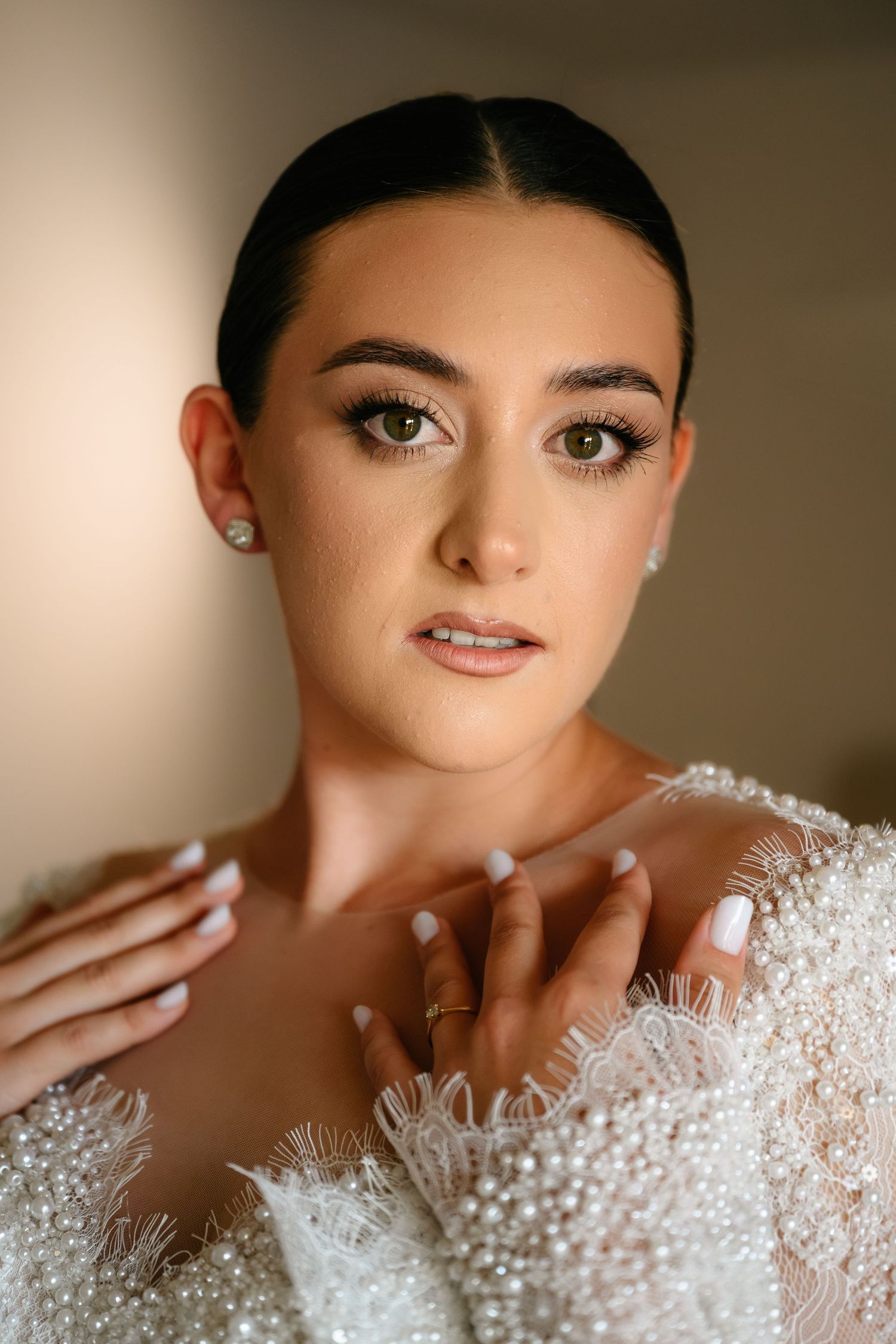 Elegant bride with sleek updo, dramatic eye makeup, and pearl-embellished white gown poses gracefully for bridal portrait.