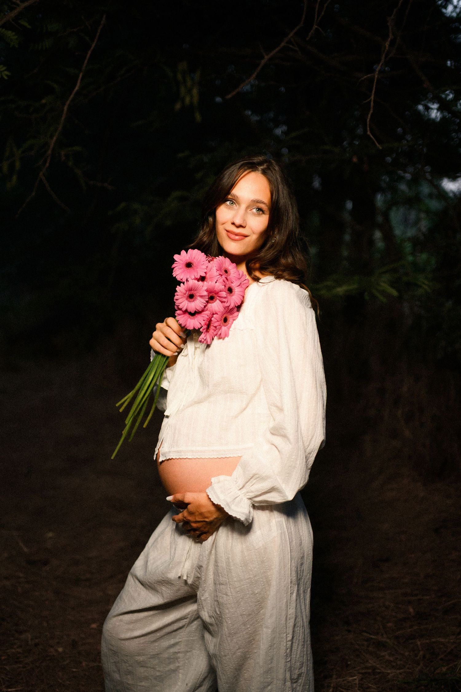 Pregnant woman in white linen outfit holding pink gerbera daisies bouquet outdoors in forest setting.