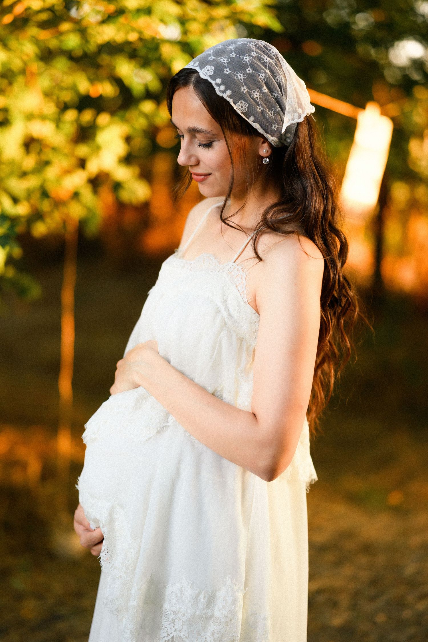 Pregnant woman in white lace dress and lace headband gently holding baby bump in warm autumn outdoor setting.